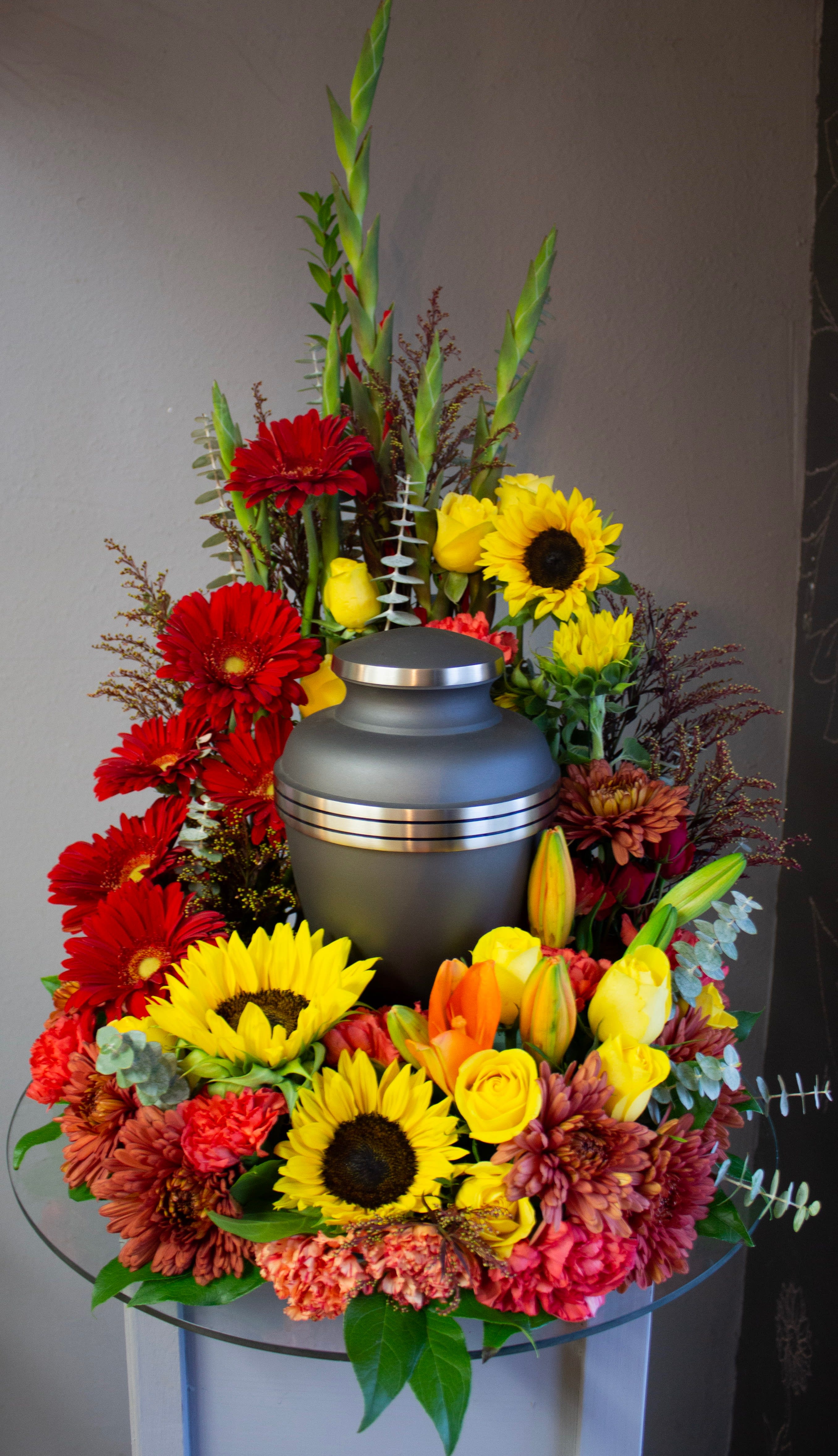 FUNERAL URN WREATH - Sunflowers &amp; Love 95 - This is a beautiful option for a funeral service with warm colors. This open ring wreath will be designed with a 10.5” ring form with the urn resting in the middle. This piece features reds and yellow flowers including gladiolas, roses, gerbera daisies, sunflowers, and lilies for a look of warmth and love. This is a stunning option for a cremation service to memorialize your loved one in beauty.  **This is a piece for a funeral or memorial service. Please place the order at least 2 days prior to the service to guarantee a certain arrangement. Anything sooner and we cannot guarantee it will match the picture. These pieces can be customized to the color of your choice. The flowers may vary depending on availability of the flowers, special requests and quality of the flowers we have in stock. If you have a special request please add it to our special instructions when placing your order. If you have any questions please don’t hesitate to call. 714 289 1123**