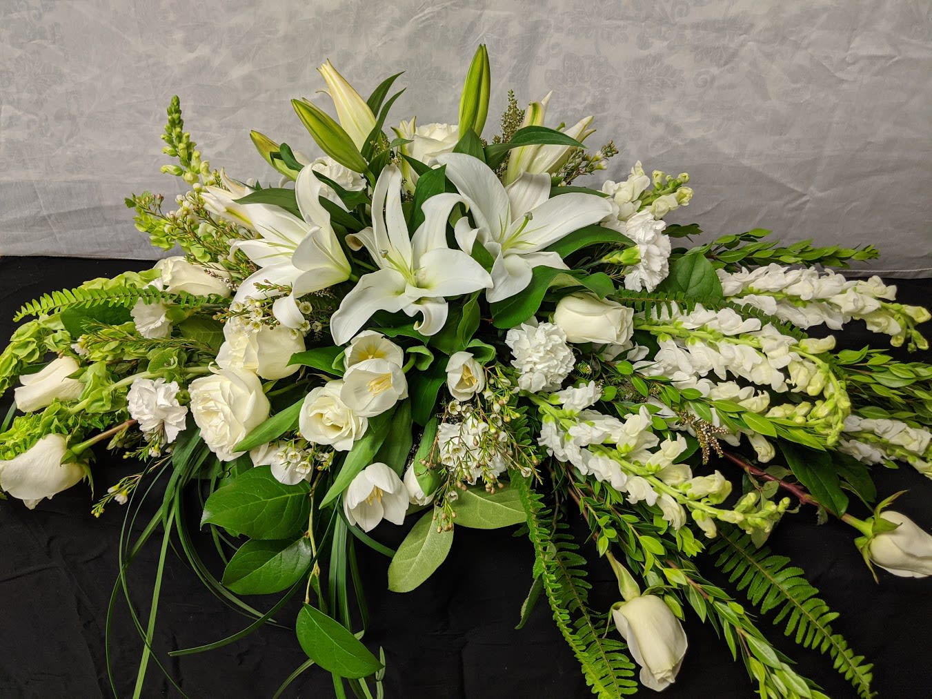 FUNERAL HALF CASKET SPRAY - Peaceful Goodbyes 1 - This is a wonderful option for a funeral service or church. This half casket will be designed with a saddle that rests approximately 4’ across the casket. This lovely mix of white roses, lilies, tulips and bells of ireland is a beautiful piece.   **This is a piece for a funeral or memorial service. Please place the order at least 2 days prior to the service to guarantee a certain arrangement. Anything sooner and we cannot guarantee it will match the picture. These pieces can be customized to the color of your choice. The flowers may vary depending on availability of the flowers, special requests and quality of the flowers we have in stock. If you have a special request please add it to our special instructions when placing your order. If you have any questions please don’t hesitate to call. 714 289 1123**