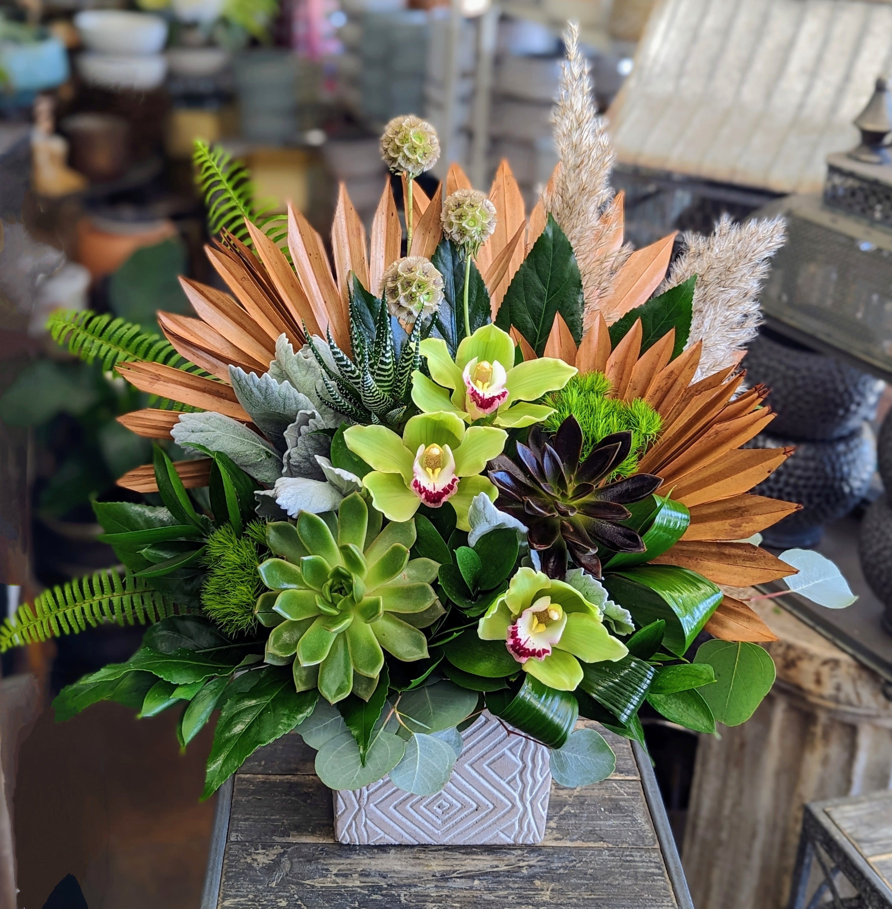 Olive - Olive by Windmill Floral Expressions in Las Vegas is a bold botanical sculpture—modern, architectural, and rich in texture. Designed to make a statement, this striking arrangement evokes the soul of the desert with a sophisticated, editorial edge.  At its core, terracotta-hued dried palm fans radiate outward like a sunburst, forming a dramatic backdrop for a palette of sculptural greens and earthen tones. Vibrant green cymbidium orchids, with deep crimson-striped throats, offer a luxurious, exotic touch, while succulents—including echeveria and dark-toned aeonium—anchor the design with grounded elegance.  Dusty miller, zebra haworthia, sword fern, and scabiosa pods add layers of movement and intrigue, while feathery pampas grass plumes rise gently above, bringing balance and softness to the arrangement’s structured form.  Housed in a patterned ceramic container, Olive feels like a modern desert garden translated into floral art—wild yet intentional, organic yet refined.  Perfect for contemporary homes, office settings, or as a luxury gift for plant and design lovers.  Available for same-day delivery in Las Vegas and Henderson.