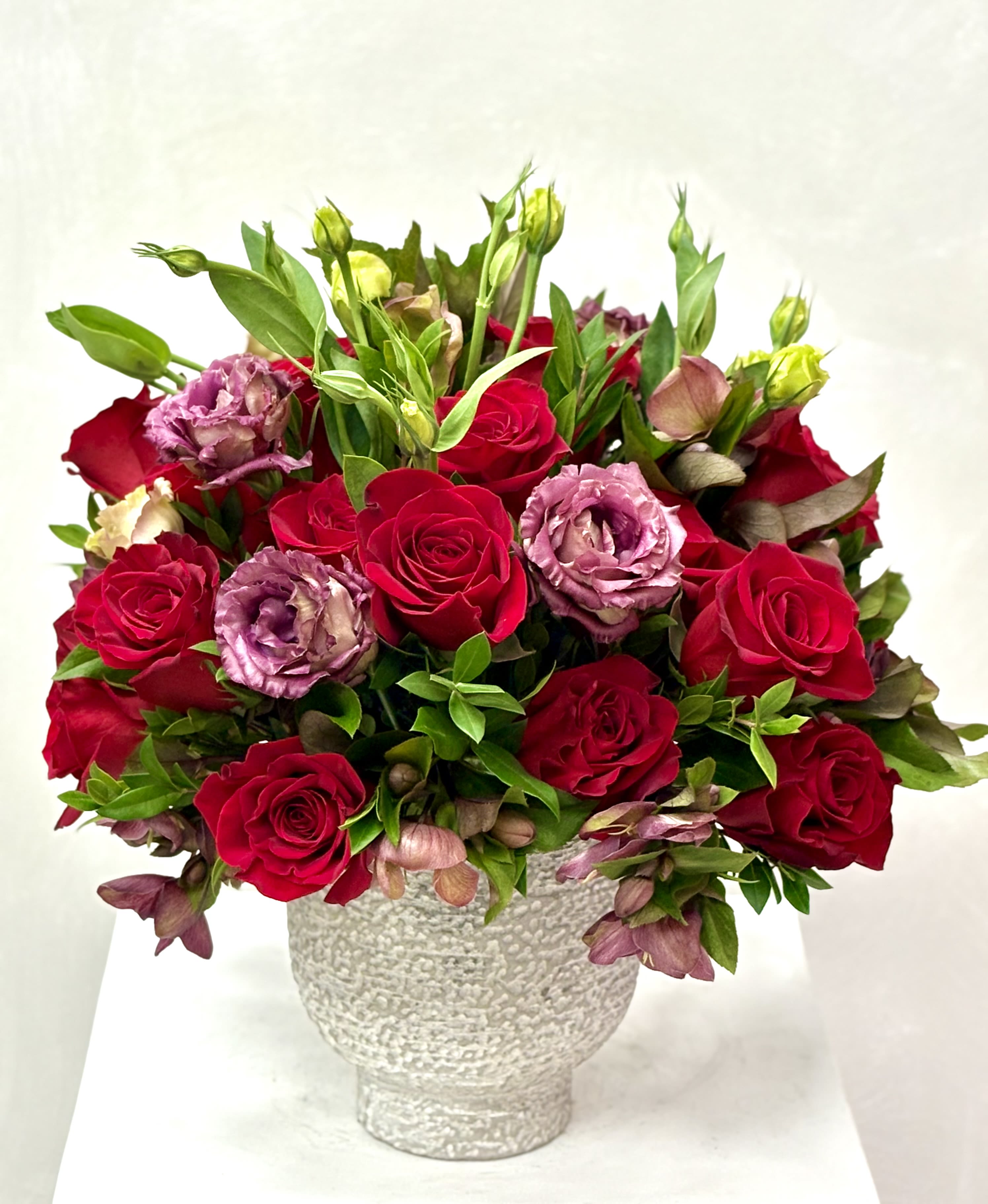 Crimson Plum Elegance By VLM Flowers - A rich and sophisticated arrangement featuring velvety red roses paired with soft plum-toned lisianthus and fresh green accents. Designed in a textured  vase, this bouquet blends deep romance with modern elegance. Perfect for anniversaries, heartfelt celebrations, or making a bold and refined statement.