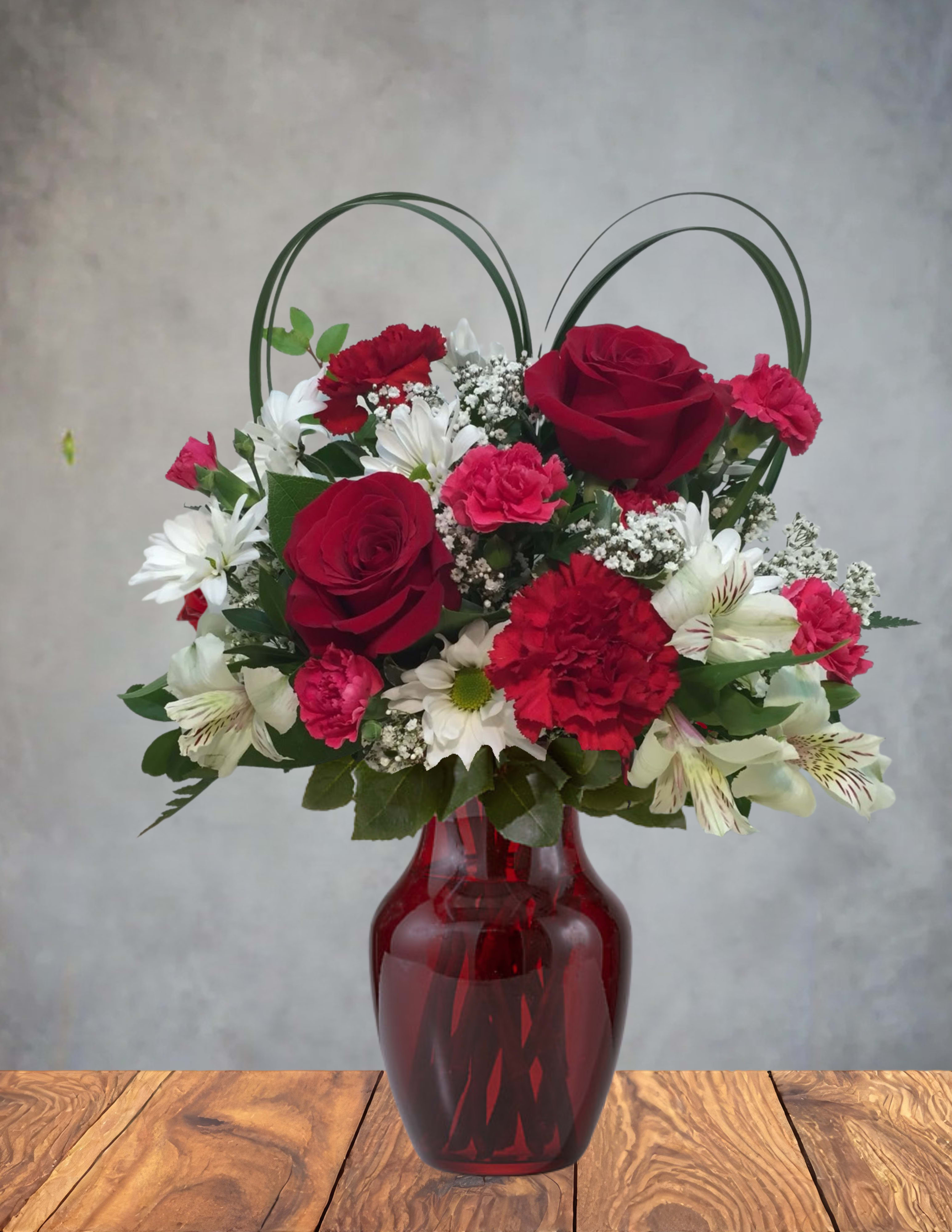 Red velvet romance  - Red Velvet Romance A lush swirl of passion and softness, this arrangement blends deep red blooms with delicate pinks and crisp whites, all gathered in a rich ruby vase that feels as indulgent as its name. The heart-shaped greenery rises above the flowers like a quiet promise, giving the whole piece a warm, intimate glow. It’s the kind of bouquet that doesn’t just say “I love you”—it lingers, like velvet on the skin and sweetness on the tongue.