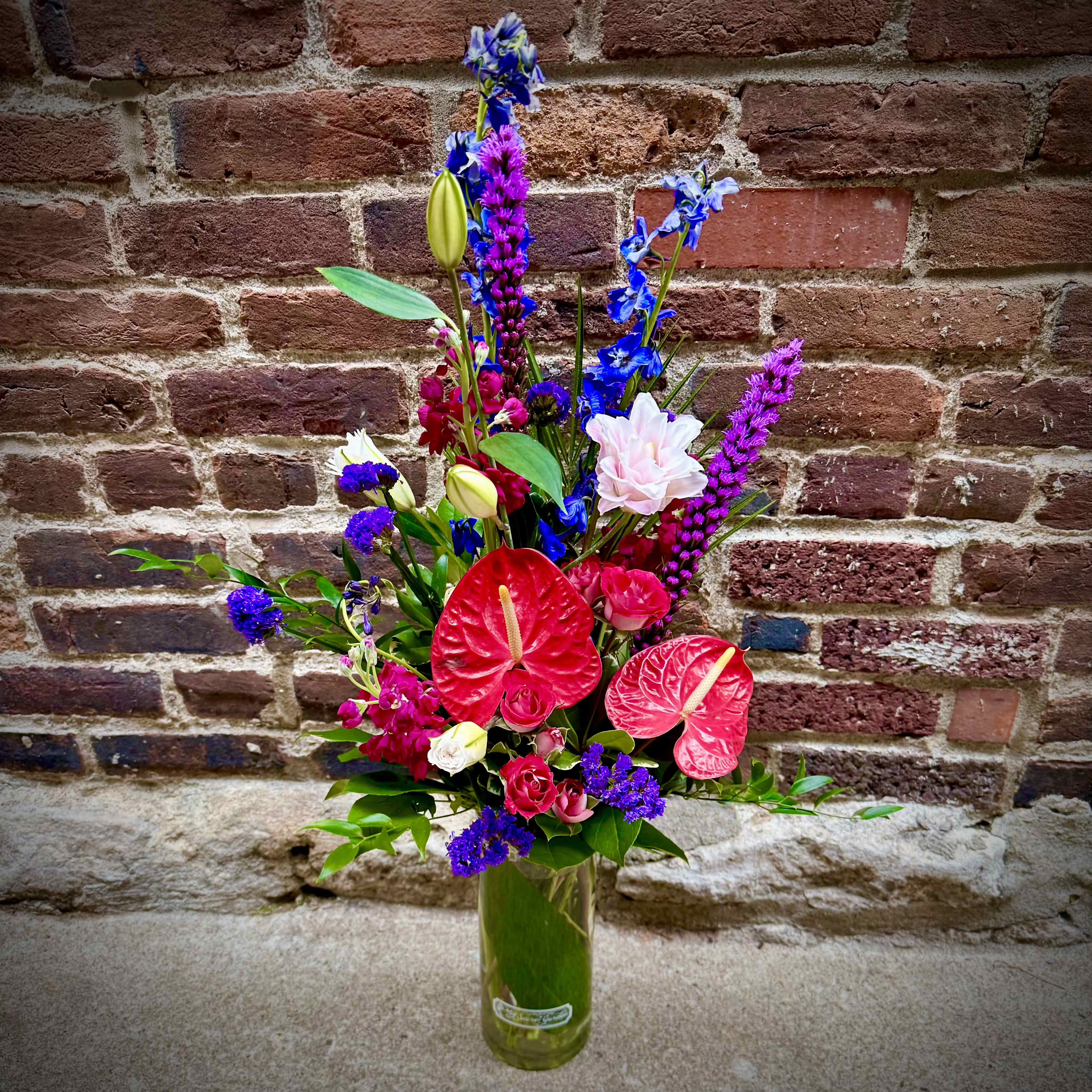 Touch of Vibrance  - "Touch of Vibrance" is a beautiful and unique floral design that is sure to bring sparkle to any occasion! Including tropical Anthurium, Liatris, Delphinium , tulip Lilies, spray roses, and stock in colors of magenta, blue, purple and white, this design is perfect for any office or home space