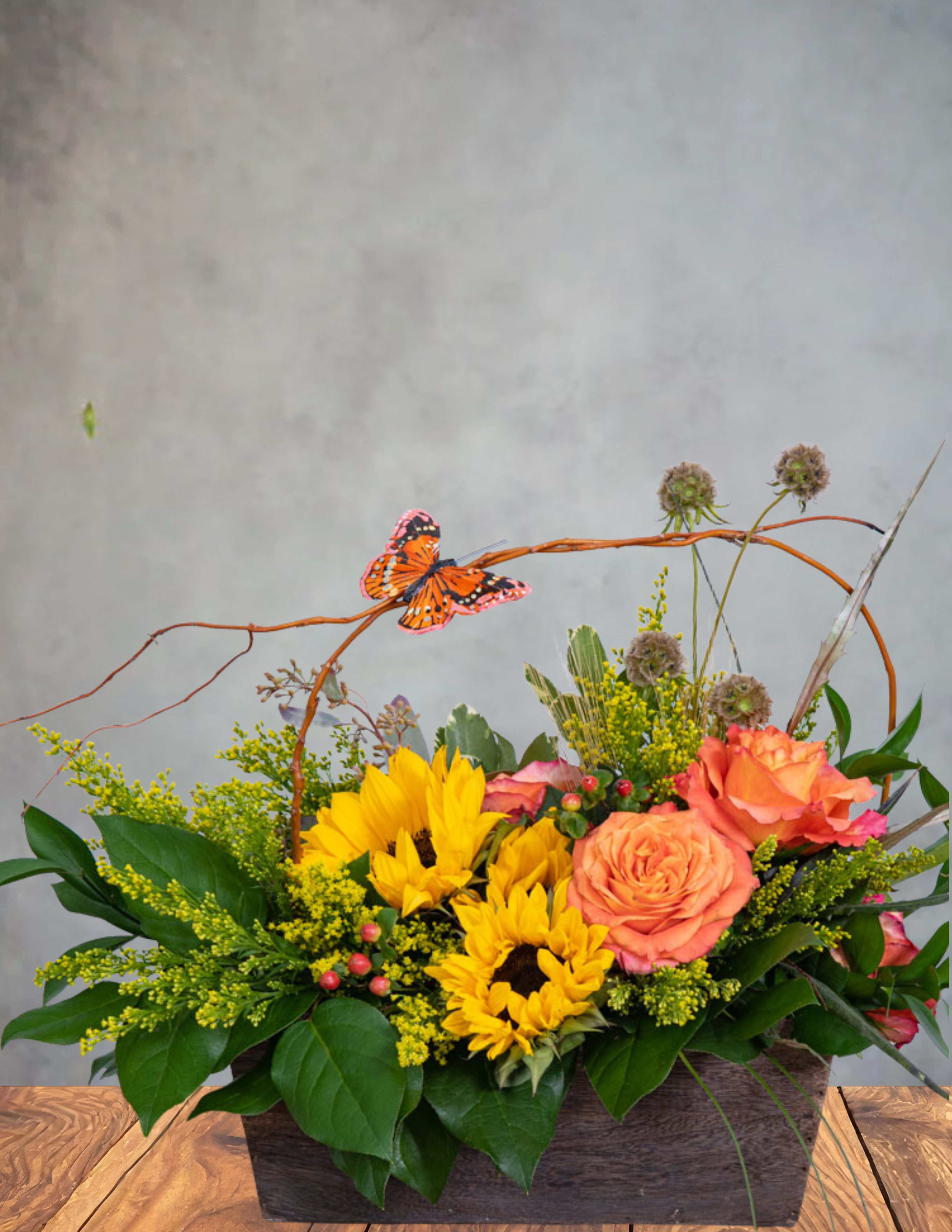 Butterfly sunrise - A burst of golden morning light captured in blooms. Butterfly Sunrise blends radiant sunflowers, glowing orange roses, and lush seasonal textures into a design that feels like the very first warm rays of the day. A graceful butterfly rests atop a natural twig arch, adding a touch of wonder and movement—as if it just fluttered in to greet the moment.