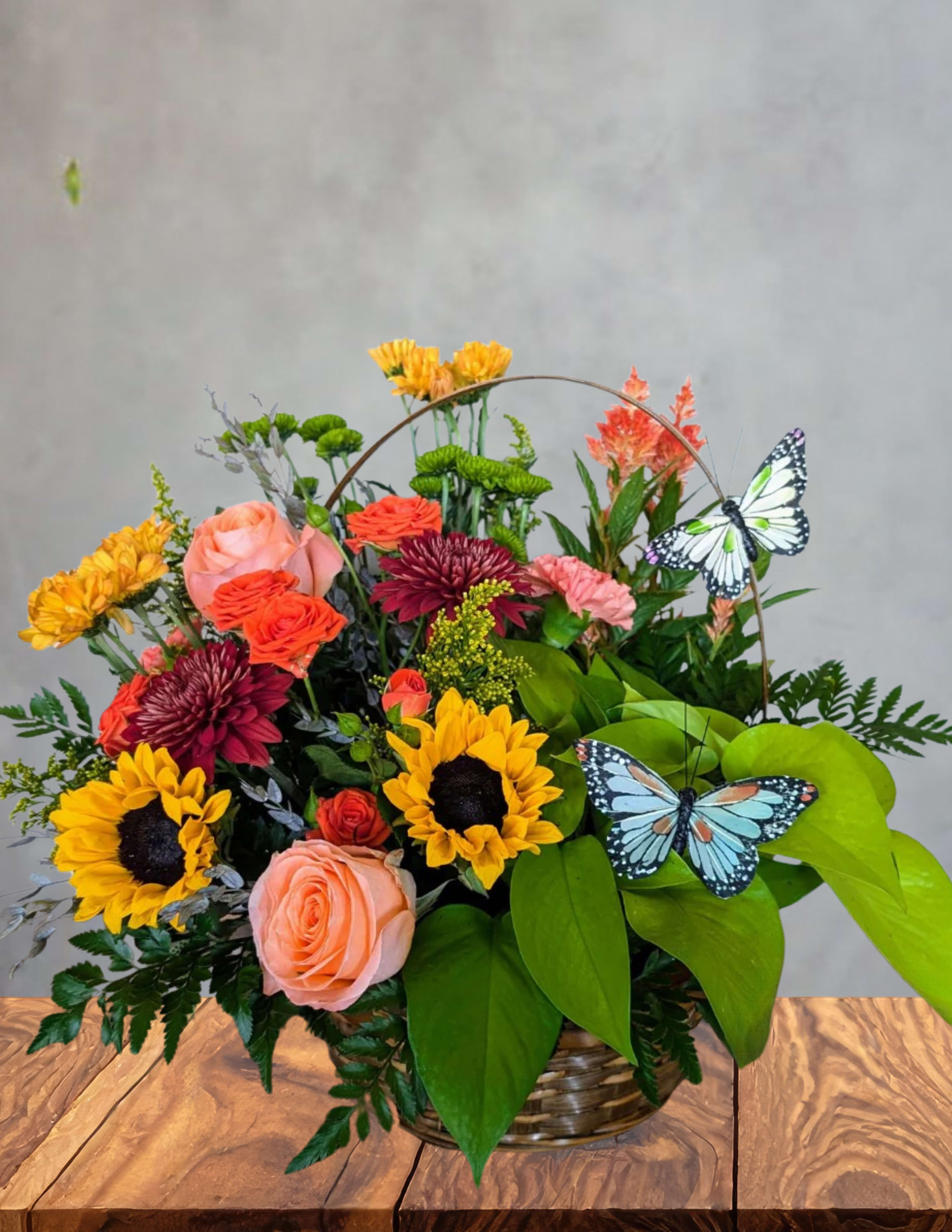 Butterfly meadow - A cheerful stroll through a sun‑kissed garden, gathered in a basket. Butterfly Meadow overflows with vibrant blooms — radiant sunflowers, playful mums, rosy accents, and lush greenery — creating a tapestry of color and texture that feels alive with movement. Two delicate butterflies rest among the petals, adding a whimsical touch as if they’ve just fluttered in to explore the blossoms.  Bright, joyful, and full of natural charm, this arrangement is perfect for celebrating life’s sweet moments or bringing a burst of meadow magic into someone’s day.