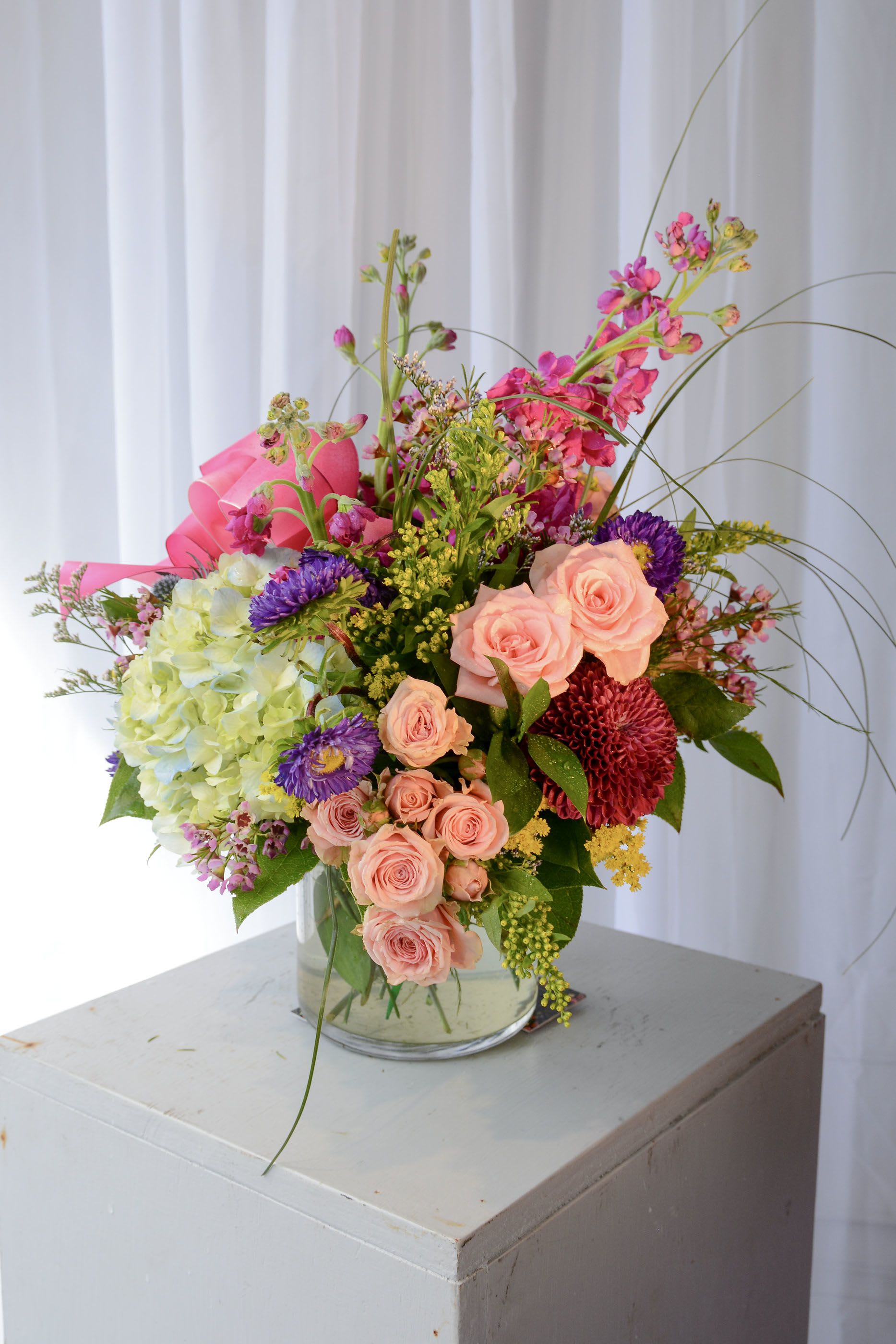 Isabella -  A French-styled flower arrangement features lush hydrangeas as the focal point, surrounded by ruffled stock flowers for texture and fragrance. Cheerful mums add vibrant color, while delicate caspia sprigs bring lightness and movement. Velvety lisianthus in shades of lavender and white enriches the elegance, and an assortment of luxury greens frames the bouquet. To finish, a beautiful satin ribbon ties the arrangement together, adding a touch of romance and sophistication to this garden-inspired display.