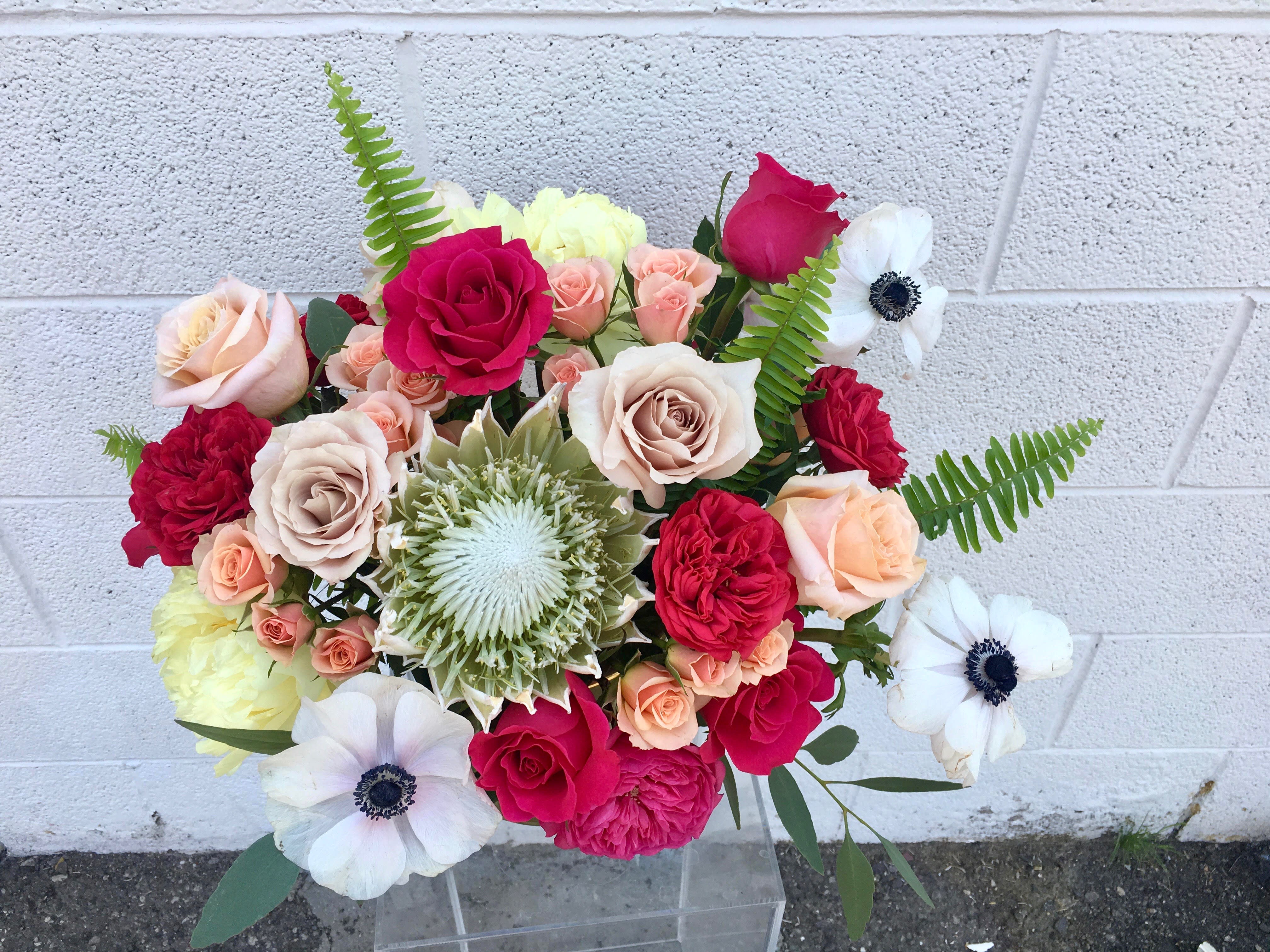 Florist's Picks - Out Of Office - DUE TO COVID-19, AVAILABILITY MAY VARY. PLEASE CALL FOR DETAILS. ALL ARRANGEMENTS WILL BE DESIGNER'S CHOICE. DELIVERY DATE AVAILABILITY IS SUBJECT TO CHANGE This vibrant and textured arrangement is designed using seasonal bold flowers. This may include mixed hot pink, light yellow, white, and peach blooms such as roses, anemones, king protea, garden roses, spray roses, touches of greenery, and other beautifully  toned elements from the Miss Daisy cooler!