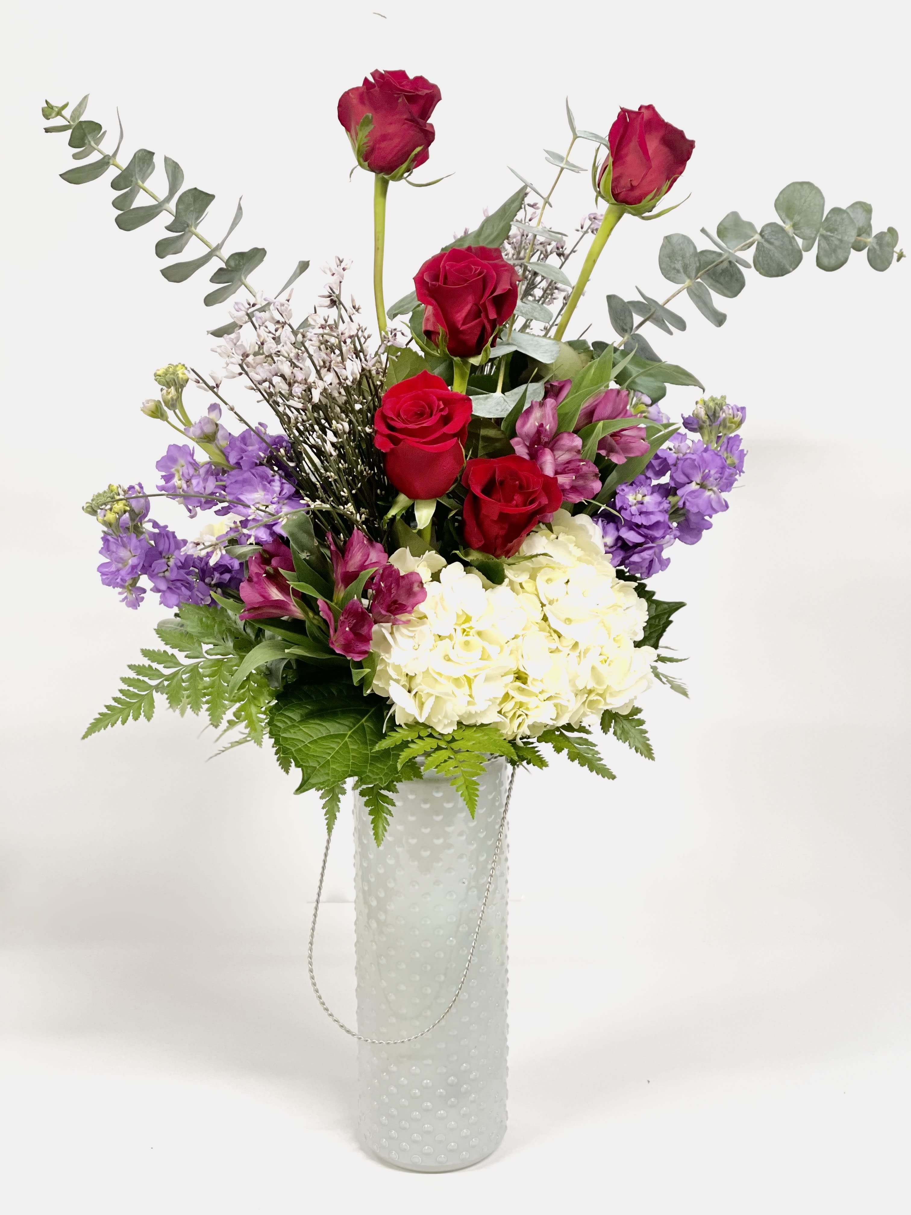 Aurora - This sky scraping beauty reaches for the stars! Designed in a tall white textured glass vase, this beauty includes red roses, lavender stock, alstroemerias, hydrangeas, eucalyptus and genestra!