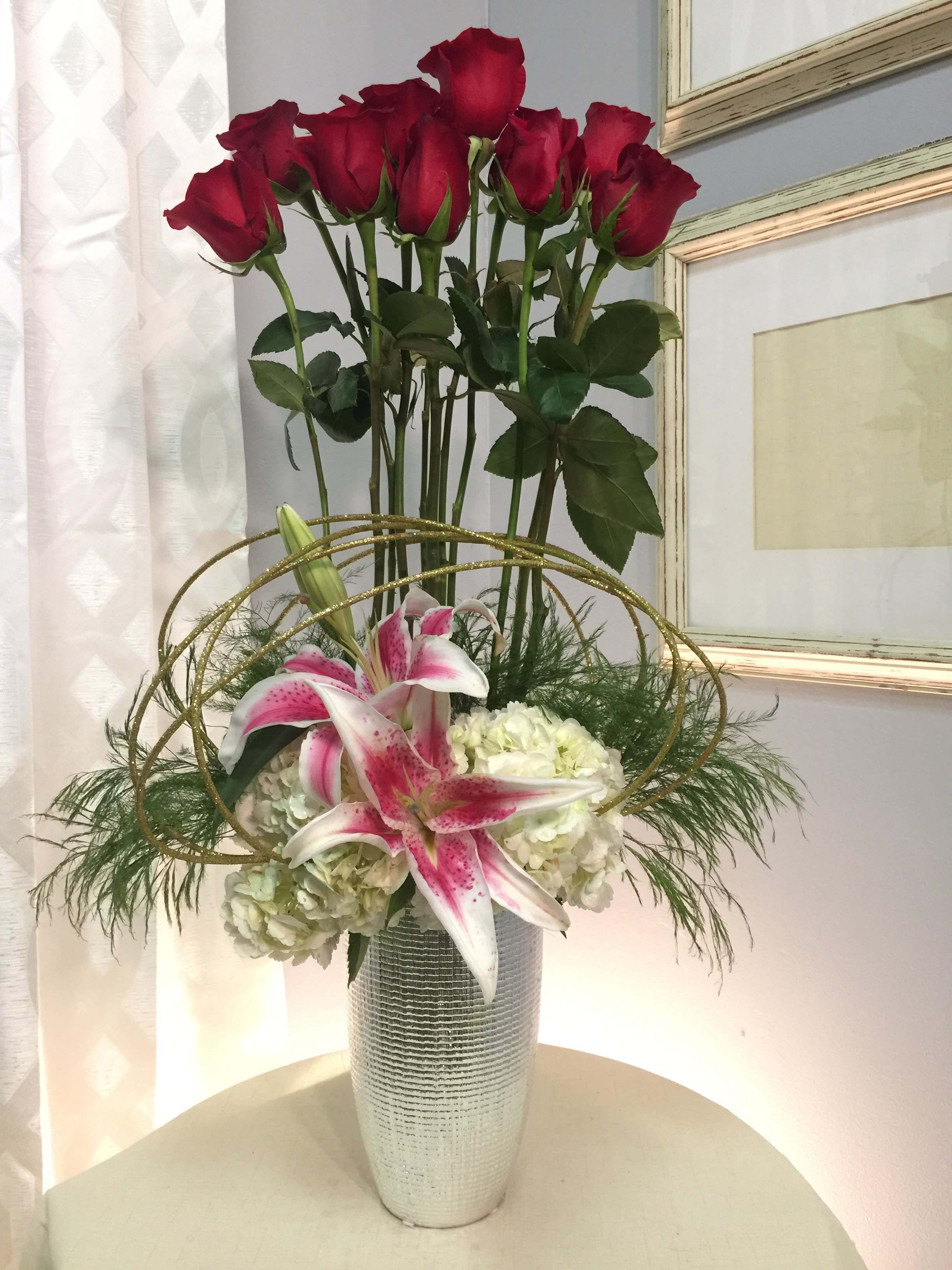 Lily Louise - A dozen red roses standing tall above a bed of white hydrangeas with stargazer lilies and glitter accents. Arranged in a keepsake silver vase.