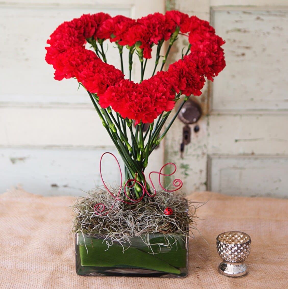 Valentina - This high styled pretty lady is made of red carnations shaped in a heart for that special someone. Accented with tropical leaves, and a script "Love".