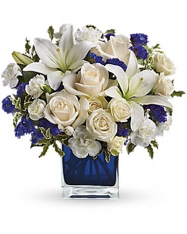 Sapphire Skies Bouquet - Send someone a bit of heaven with this beautiful bouquet. Luxurious crÃ¨me roses and pure white lilies paint a peaceful picture inside a sapphire blue cube. CrÃ¨me roses, white asiatic lilies and white miniature carnations are mixed with bursts of purple statice and green pitta negra. Delivered in a glass Cube. 