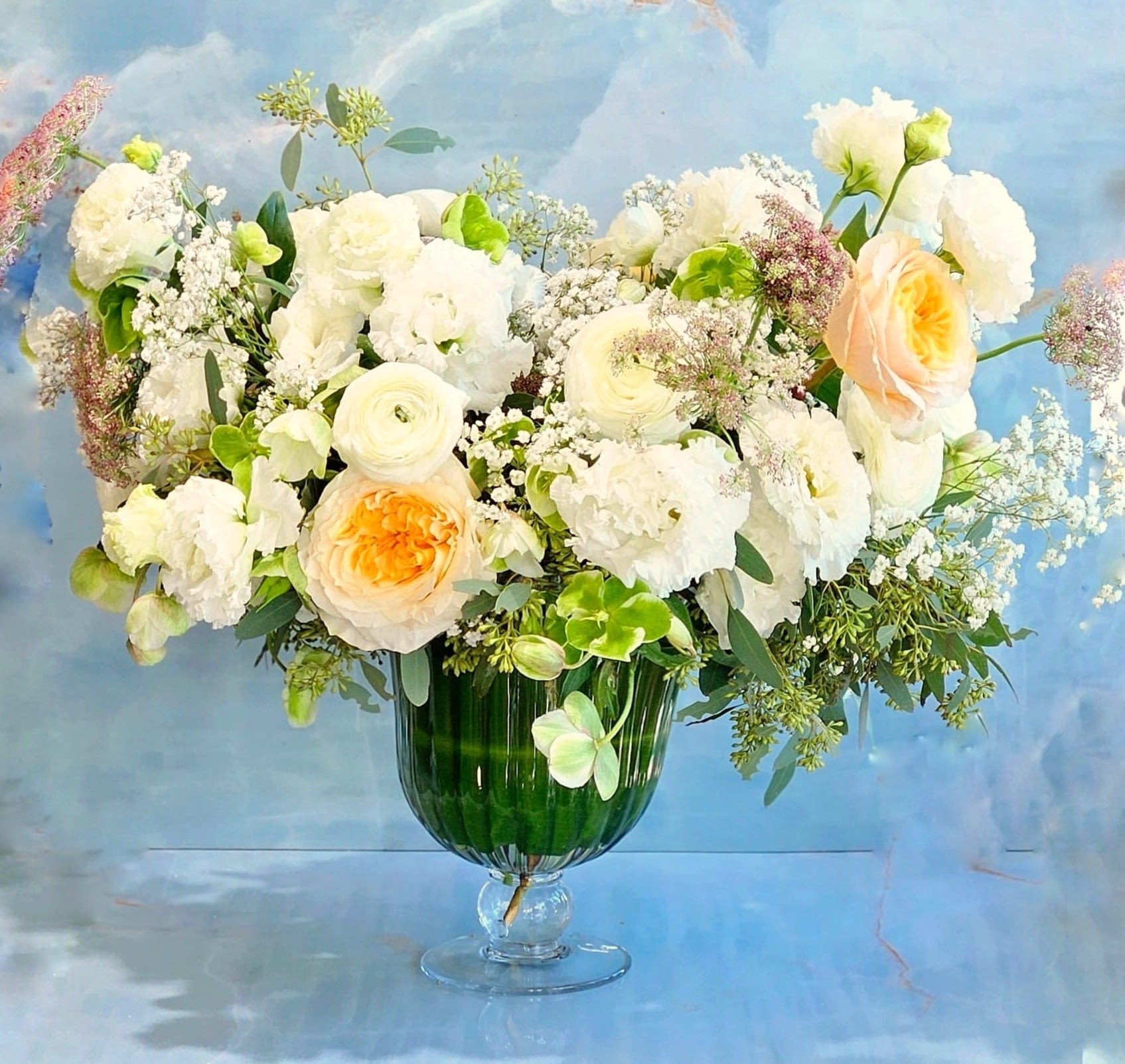 French Riviera  - This arrangement feels soft, airy, and quietly romantic, with a touch of timeless elegance.  Blush garden roses take center stage, their ruffled petals layered among creamy whites and fresh green accents. Delicate textures—like spray blooms and light filler—create movement and depth, giving the piece an effortless, garden-gathered feel. The palette stays gentle and refined, with warm blush tones balanced by crisp whites and natural greenery, all arranged loosely in a classic clear vessel.  Overall, it’s graceful and luminous, perfect for someone who loves understated beauty—romantic without being fussy, polished yet organic.