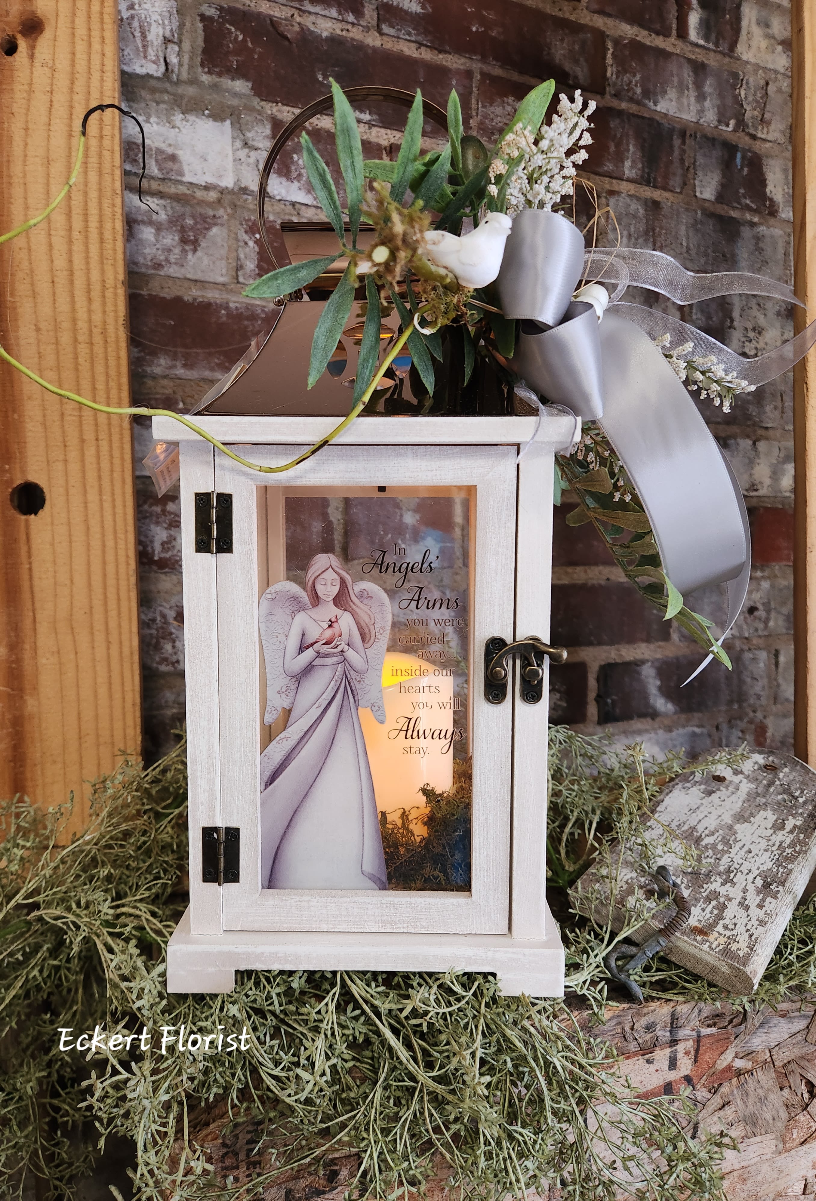 Eckert Florist's In Angels' Arms Lantern *Our local delivery only - Nice keepsake white wooden lantern with copper tin roof and sentiment on Angel motif glass. Sentiment: "In Angels Arms you were carried away...inside our hearts you will Always stay" Included is a battery operated candle. Measures approx. 6.25" Square x 15" High *OUR LOCAL DELIVERY ONLY