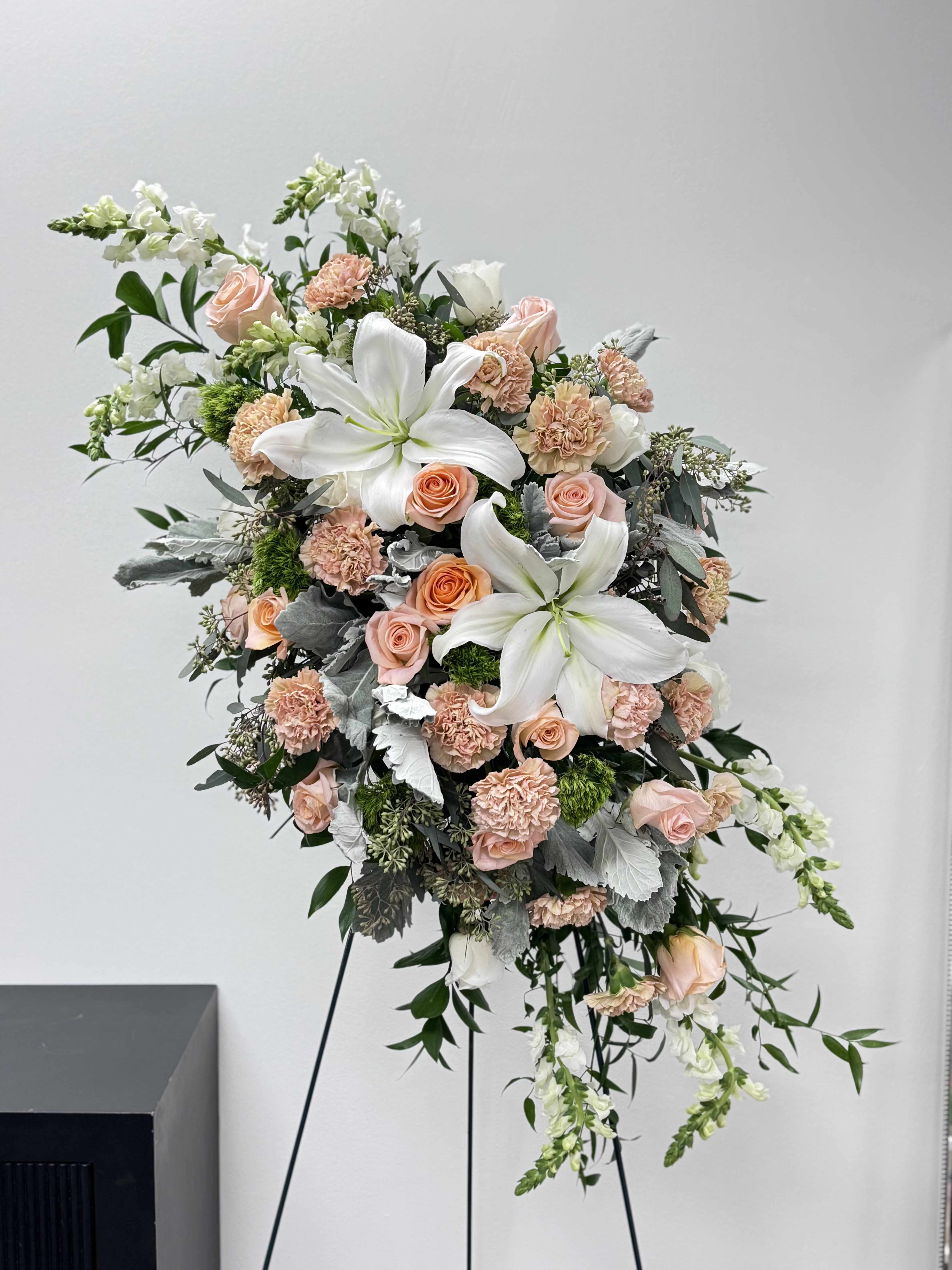 Graceful Rememerance - Honor a beautiful life with our Graceful Remembrance Standing Spray, a soft and elegant tribute designed to offer comfort and express heartfelt sympathy.  This timeless funeral spray features pristine white lilies paired with delicate blush roses and soft peach carnations, thoughtfully arranged with textured silver foliage and lush greenery. The cascading design adds gentle movement, creating a graceful presence beside the casket or at a memorial service.  The combination of creamy whites and romantic blush tones symbolizes peace, remembrance, and everlasting love — making this standing spray a meaningful way to honor someone special.  Each arrangement is handcrafted with care by our floral designers to ensure beauty, freshness, and a dignified presentation.  At Boîte de Fleurs, we understand how important it is to send something thoughtful during difficult times. We offer reliable funeral flower delivery in Phoenix, Scottsdale, and surrounding Arizona areas to ensure your tribute arrives with care and professionalism.
