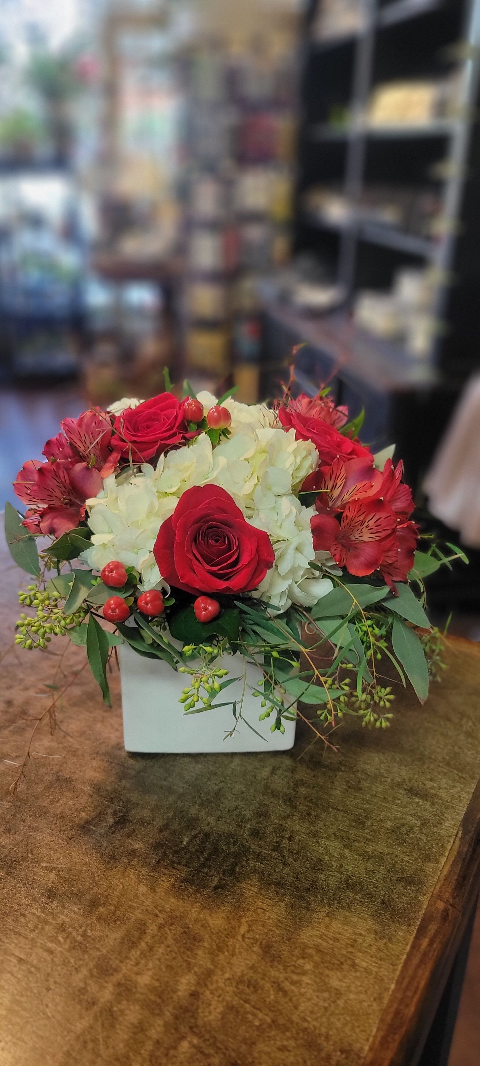 Crimson Elegance - A stunning arrangement featuring lush red roses, delicate white hydrangea, and vibrant red alstroemeria, accented with seasonal greenery and red hypericum berries. Designed in a chic white container, this arrangement exudes warmth and sophistication, making it perfect for romantic gestures, celebrations, or as a timeless centerpiece. 