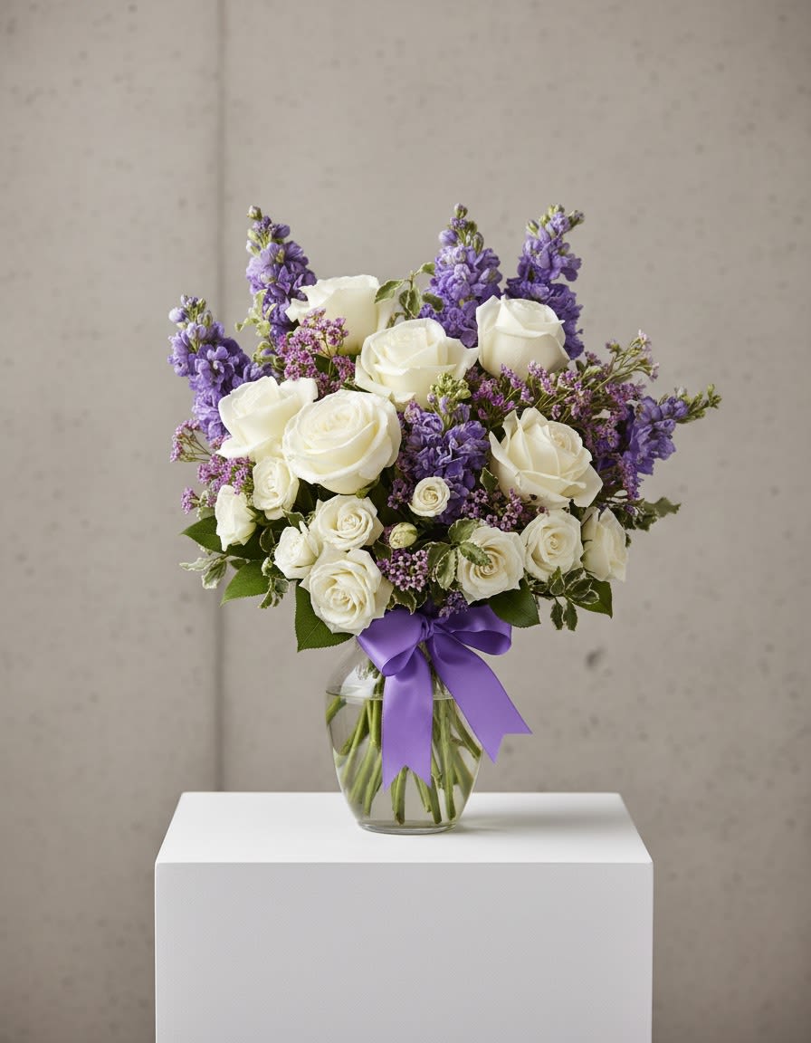 The Enchanted Cottage - As enchanting as coming across a hidden cottage in the countryside! Lush white roses and purple stock mix with delicate lavender waxflower and green pitta negra for an elegant English garden look. The decorative bouquet is finished with a pretty satin ribbon.  White roses meet lavender stock and waxflower in this lush, elegant arrangement.  Presented in a classic clear glass vase with purple satin ribbon. Orientation: One-Sided