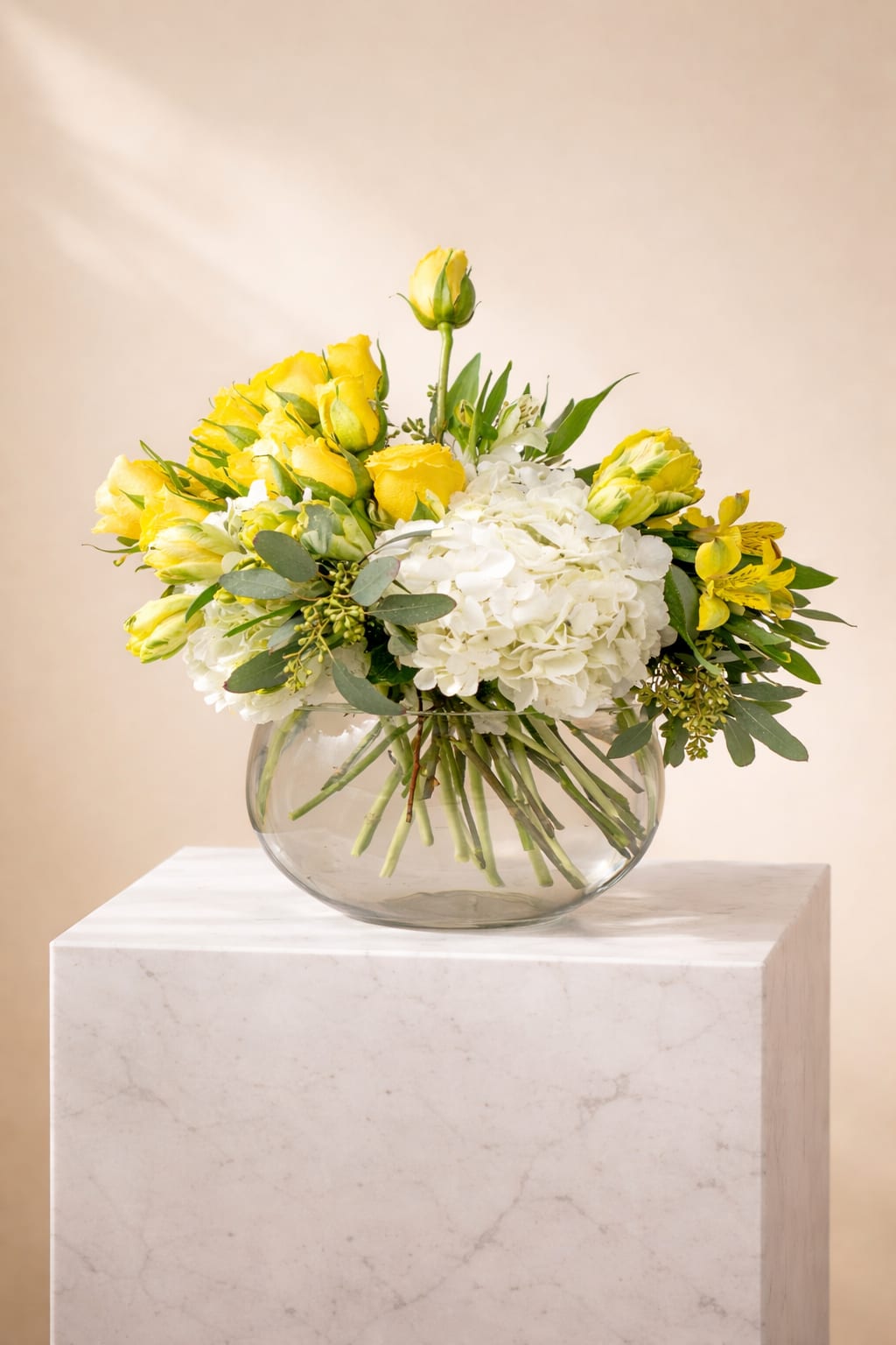 Sunlit Elegance  - A bright and refined arrangement featuring radiant yellow roses, lush white hydrangeas, and fresh seasonal blooms accented with eucalyptus greenery. Styled in a modern round glass vase, Sunlit Elegance brings warmth, light, and effortless sophistication to any space. Perfect for celebrations, thank-you gestures, housewarmings, or simply brightening someone’s day.