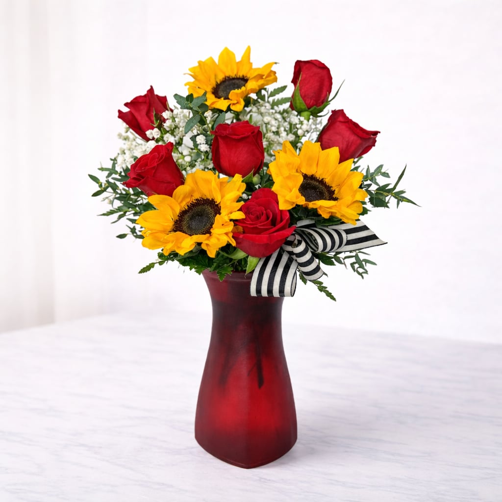 Sunlit Romance - Sunlit Romance pairs vibrant sunflowers with classic red roses, soft white accents, and fresh greenery for a bold yet cheerful design. Arranged in a rich red glass vase and finished with a our signature black-and-white ribbon, this arrangement brings warmth, love, and joy—perfect for anniversaries, celebrations, or making someone smile.