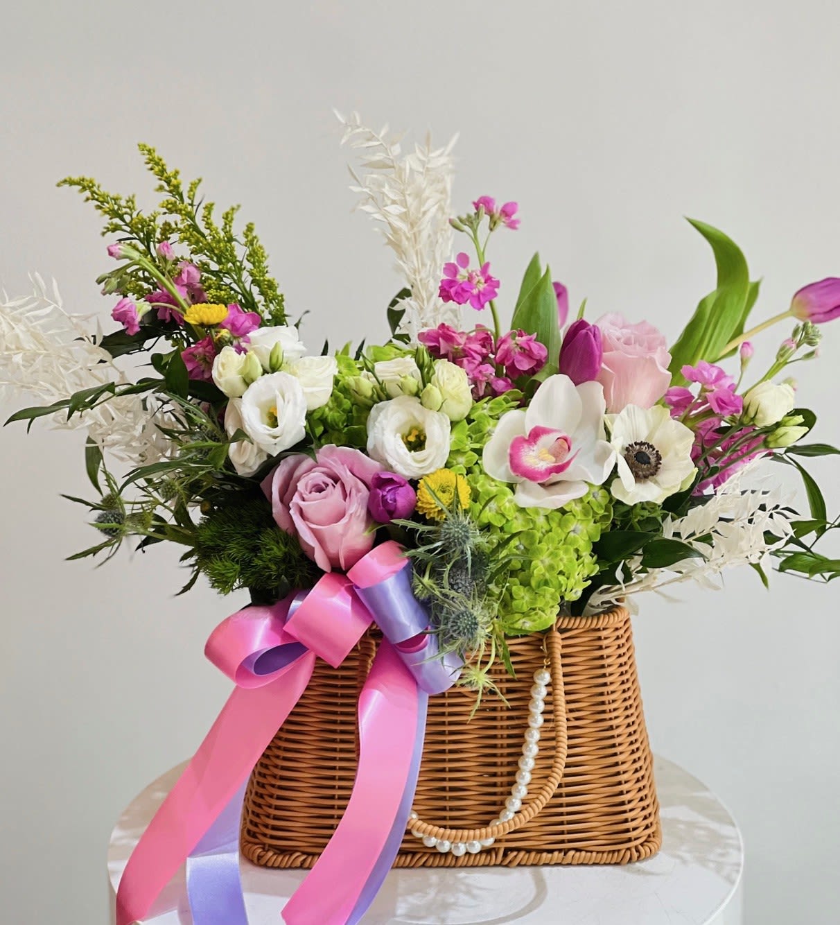 Secret Garden ™ - Set in an elegant woven basket with pearl chain and satin ribbon, this lavish arrangement overflows with extraordinary anemones, green hydrangeas, roses, tulips, lisianthus, stock, thistle, and preserved foliage. A rare and luxurious design, it blends texture, elegance, and drama into a showpiece that captivates from every angle.