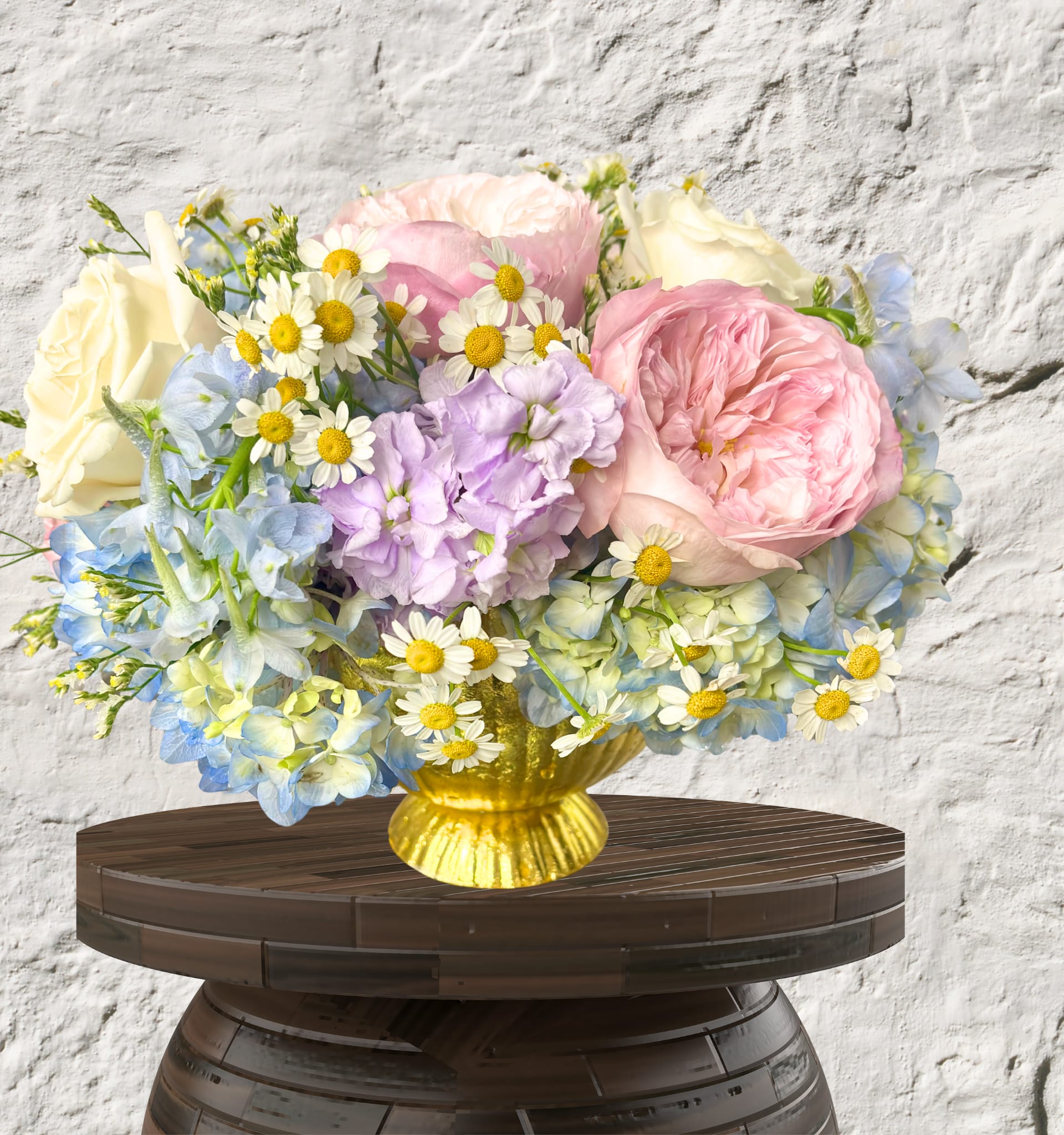 Golden Meadow Bouquet - The Golden Meadow Bouquet Sun-kissed charm meets garden elegance.  This whimsical, airy arrangement brings the brightness of a summer day into any room. Featuring lush pink Roses and ivory blooms, it is beautifully accented with sky-blue Delphinium, fragrant Lavender Stock, and cheerful Chamomile daisies.  Presented in a striking gold-toned pedestal vase, this arrangement offers a perfect balance of rustic charm and polished style.  Vibe: Joyful, breezy, and enchanting.  Key Blooms:  Roses, Chamomile, and Delphinium.  Perfect For: Birthdays, "thinking of you," or a bright kitchen centerpiece.