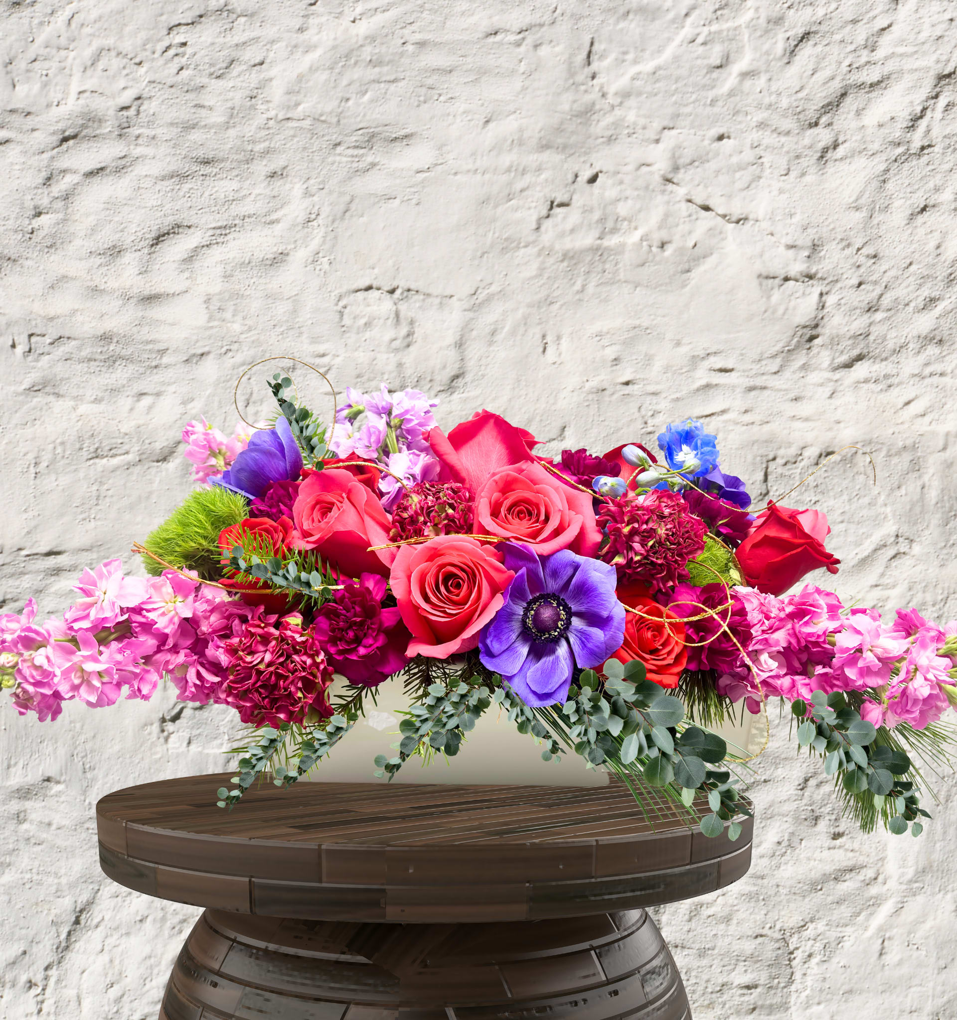 Regal Radiance - Bespoke Artistry. Jewel-Toned Splendor. Heirloom Elegance.  The Regal Radiance is a masterwork of texture and vibrant color, designed to serve as the crowning jewel of any setting. This elongated "Sovereign Statement" piece is a sprawling garden of high-contrast luxury, blending rich magentas and deep plums with striking pops of electric blue. It captures a sense of wild, sophisticated movement that flows effortlessly across a table or mantel.  The Design &amp; Florals This centerpiece is a premier example of our artisan craftsmanship, utilizing a low-profile silhouette that encourages conversation while commanding attention.  Premium Blooms: Vibrant pink and red Garden Roses anchor the design, surrounded by velvet-petaled purple Anemones and lush, ruffled carnations in deep wine hues.  Textural Accents: Billowing stems of lavender Stock provide fragrance and soft lines, while seasonal foliage, gold-accents add layers of modern, festive intrigue.  The Upscale Vessel: Presented in a sleek, elongated white ceramic boat. This clean, architectural foundation provides a crisp contrast to the overflowing jewel-toned blooms, ensuring the arrangement feels contemporary and high-end.  