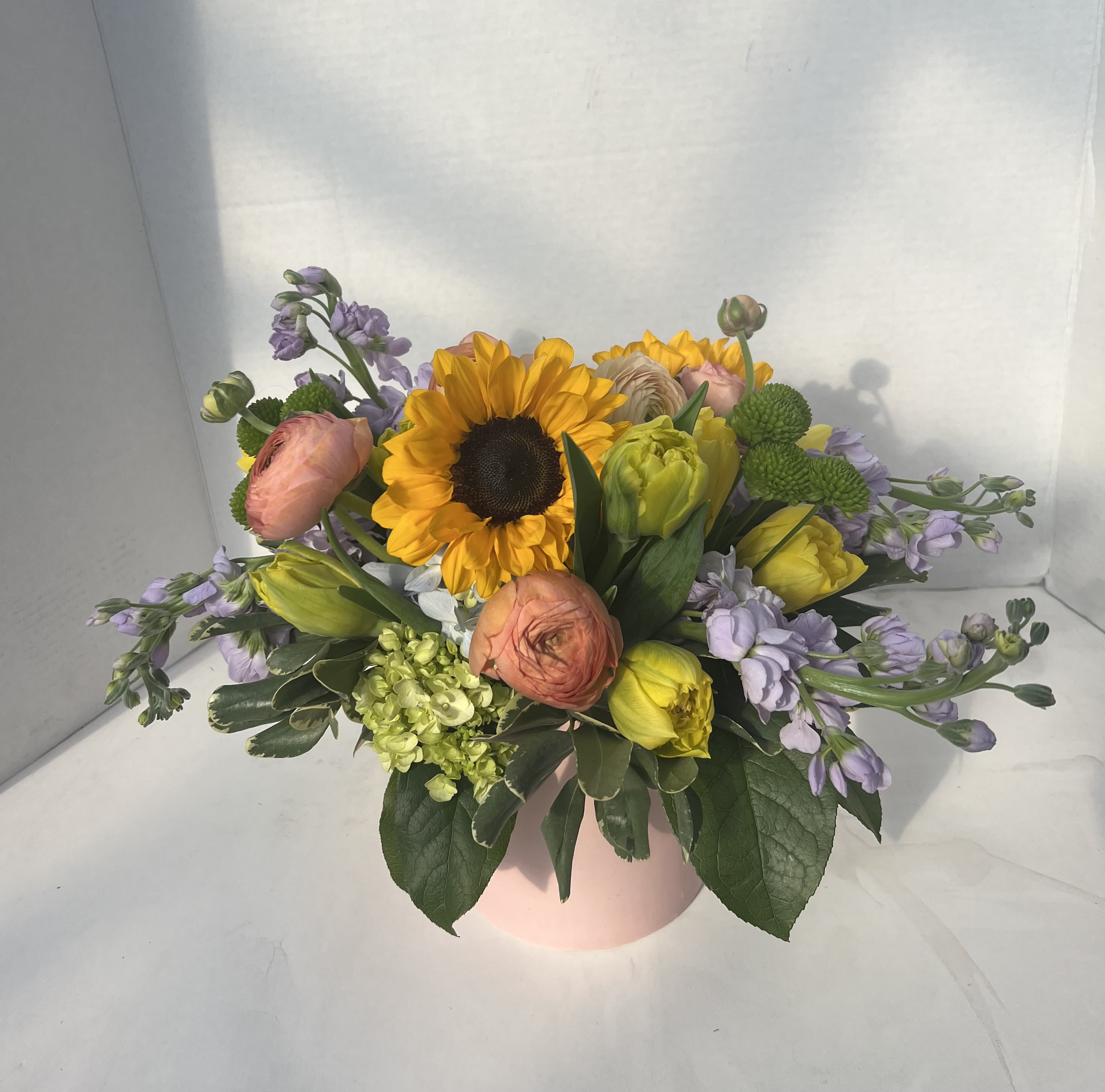 Sunny Day - Whether it’s brightening a kitchen table, celebrating a special moment, or sending a little happiness just because, Sunny Day delivers warmth, color, and joy in every petal. Sunny sunflowers, lush blue hydrangea, yellow tulips and delicately layered ranunculus blooms weave throughout the design in a pink ceramic pot.