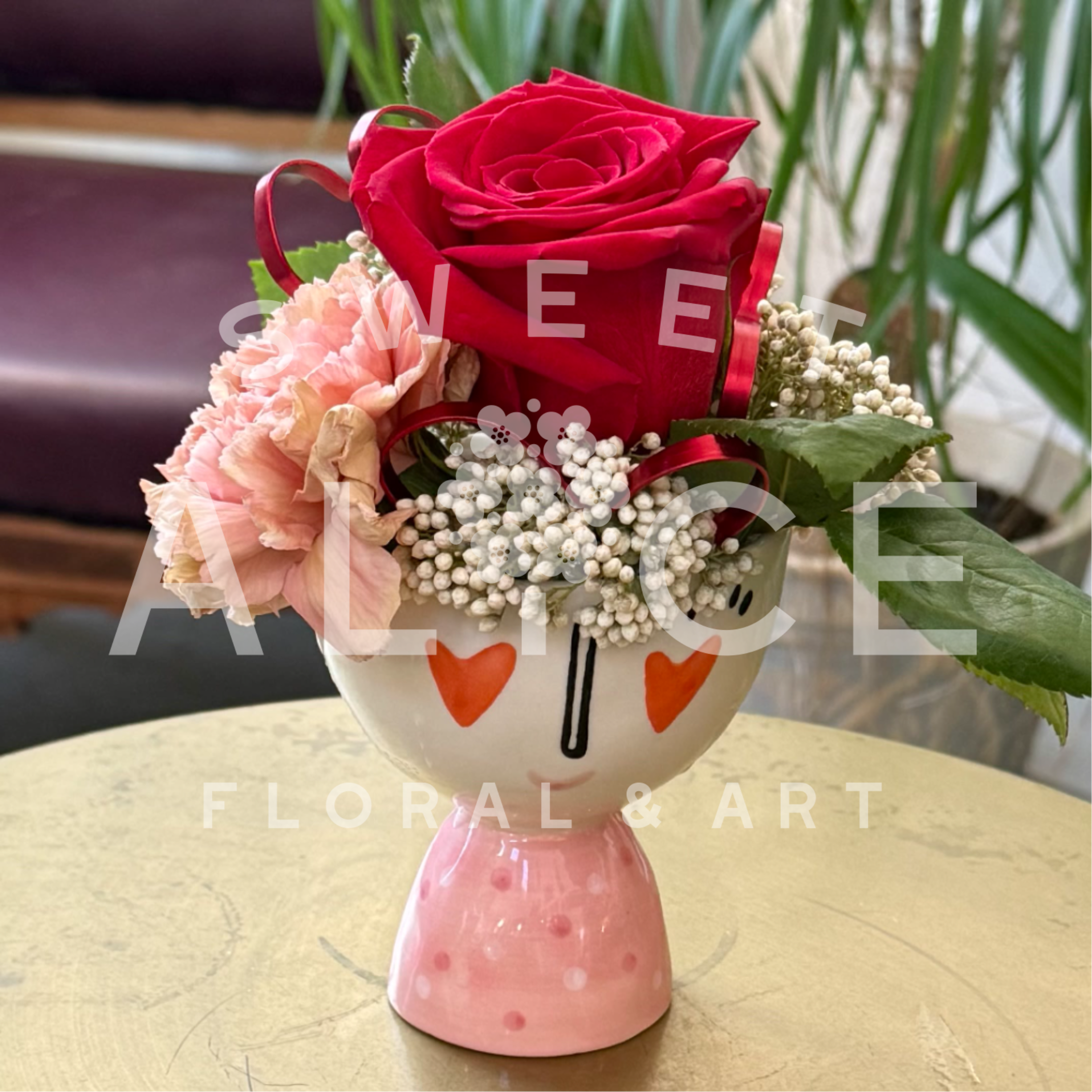 Flirt Compote - This adorable little compote is the perfect way to flirt a little. This design features a red rose, a blush carnation, and rice flower in a flirty faced cermaic footed compote. Red wire hearts dance through the composition. A truly delightful treat!