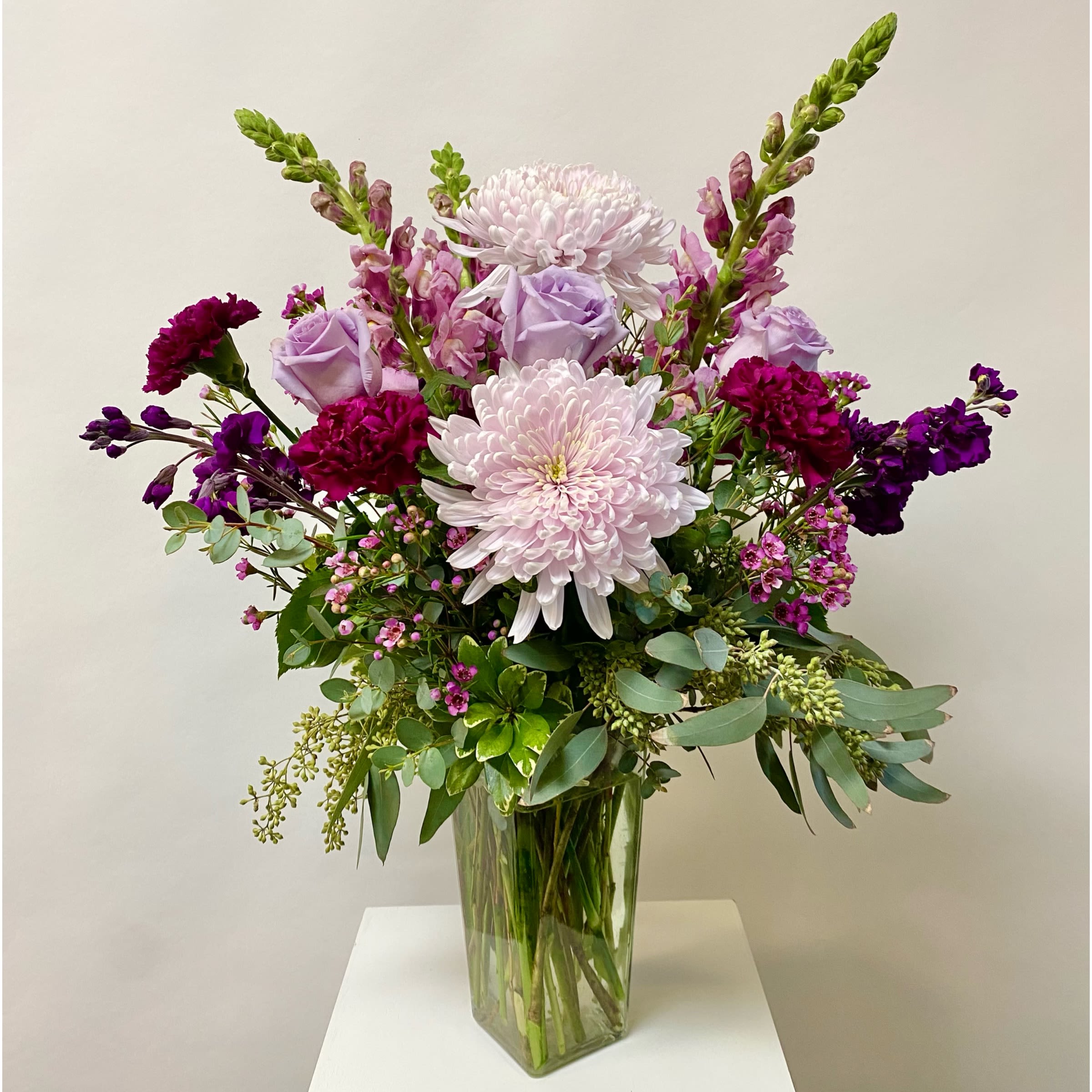 Lavish Lavender - This Tall Purple Monochrome Arrangement includes Premium Greenery, Cremone Mums, Snapdragons, Stock, Roses, Carnations, Wax Filler and Seeded Eucalyptus in a Glass Vase. Or as Similar as Possible. 