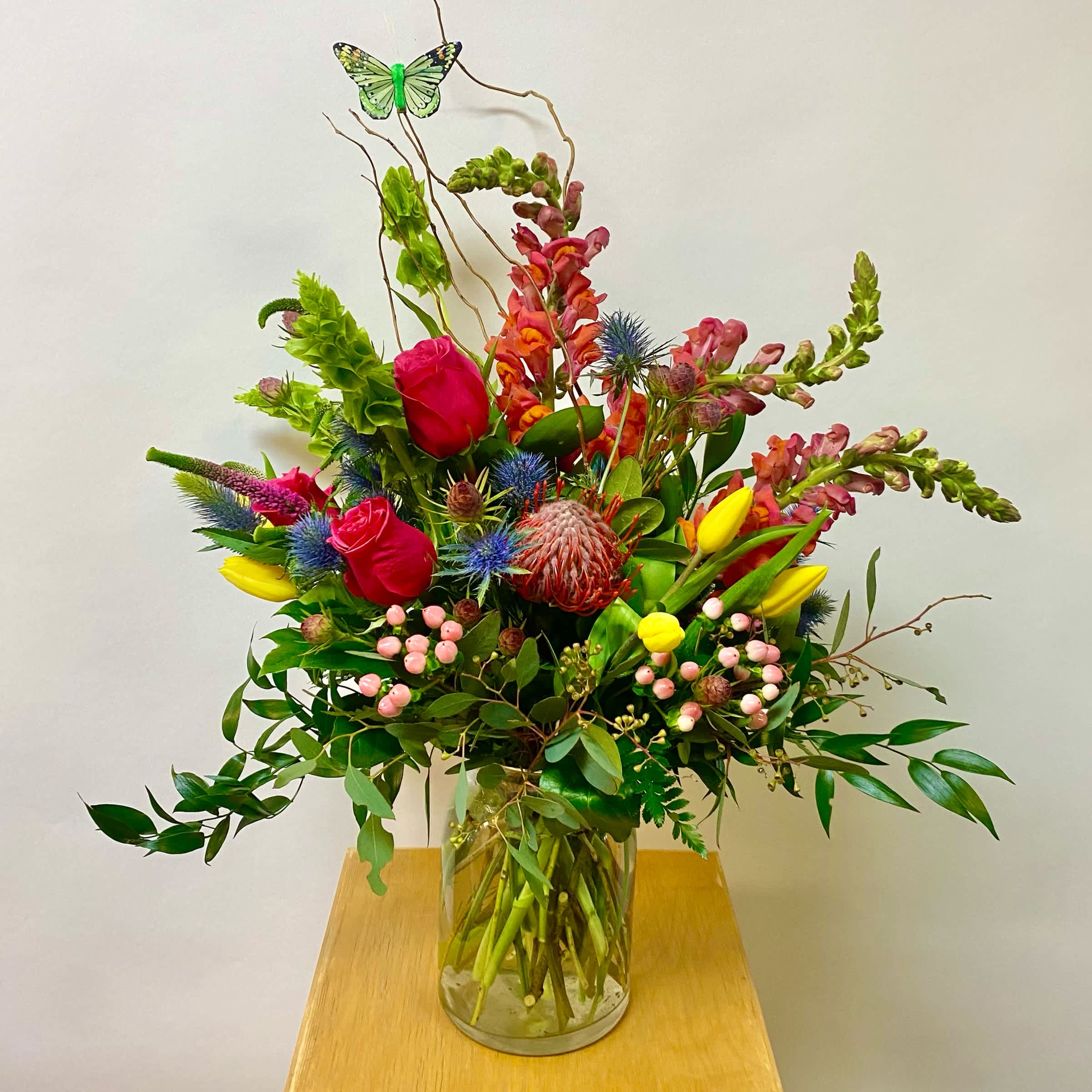 Mother's Garden - This Wild Garden Themed Arrangement includes Hot Pink Roses,  Bells Of Ireland, Yellow Tulips, Orange Snaps, Blue Thistle, Pincushion, Safari, Light Pink Berries and Curly Willow in a Medium Glass Vase and Accented with a Butterfly. Or as Similar as Possible. 