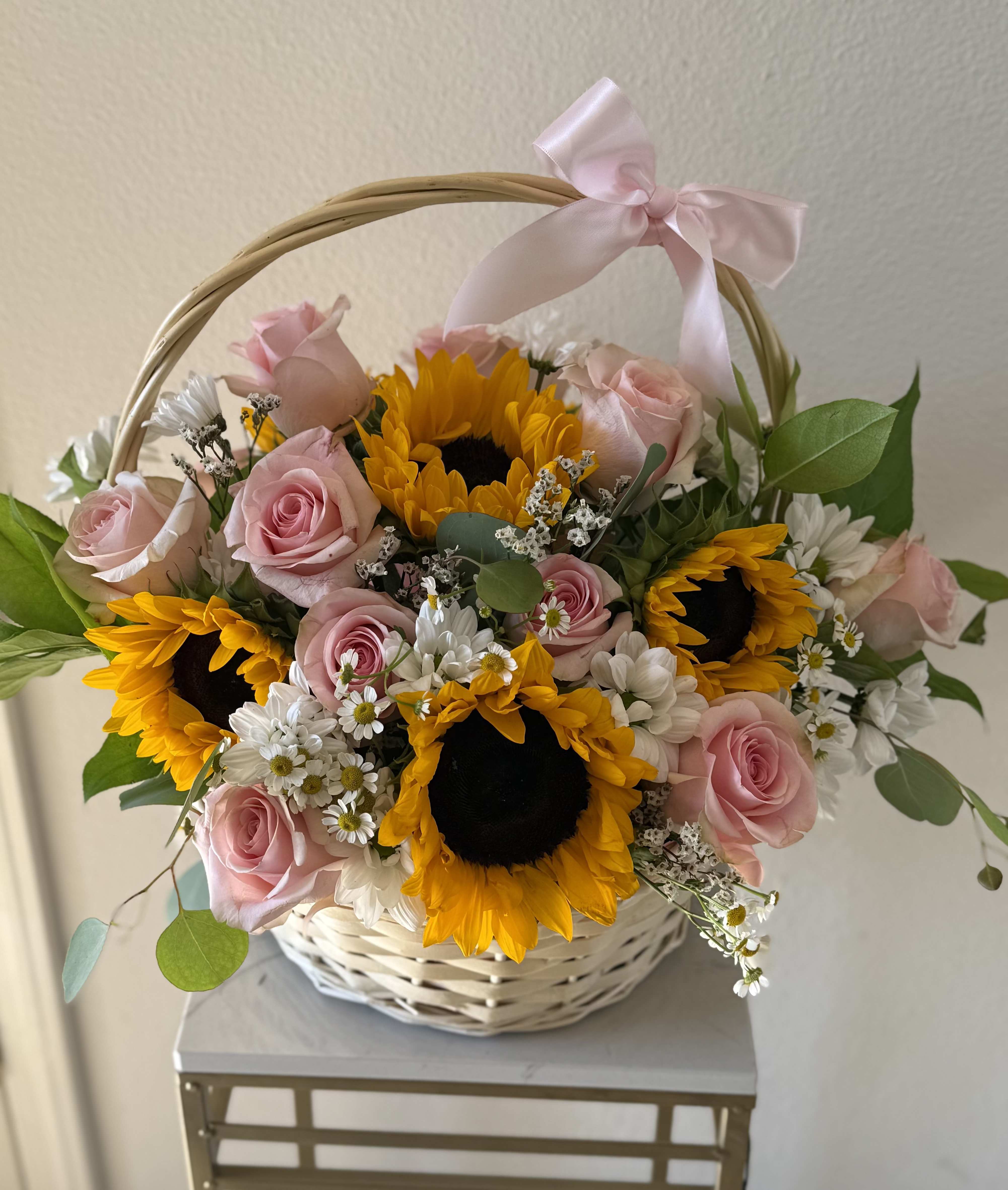 The Sunshine Basket - Bright, cheerful, and full of charm, The Sunshine Basket features radiant sunflowers, elegant pink roses, soft white daisies, and airy baby’s breath, all nestled in a classic white wicker basket and finished with a delicate satin bow. Perfect for birthdays, thank yous, or simply to brighten someone’s day.  Fresh. Uplifting. Beautiful.