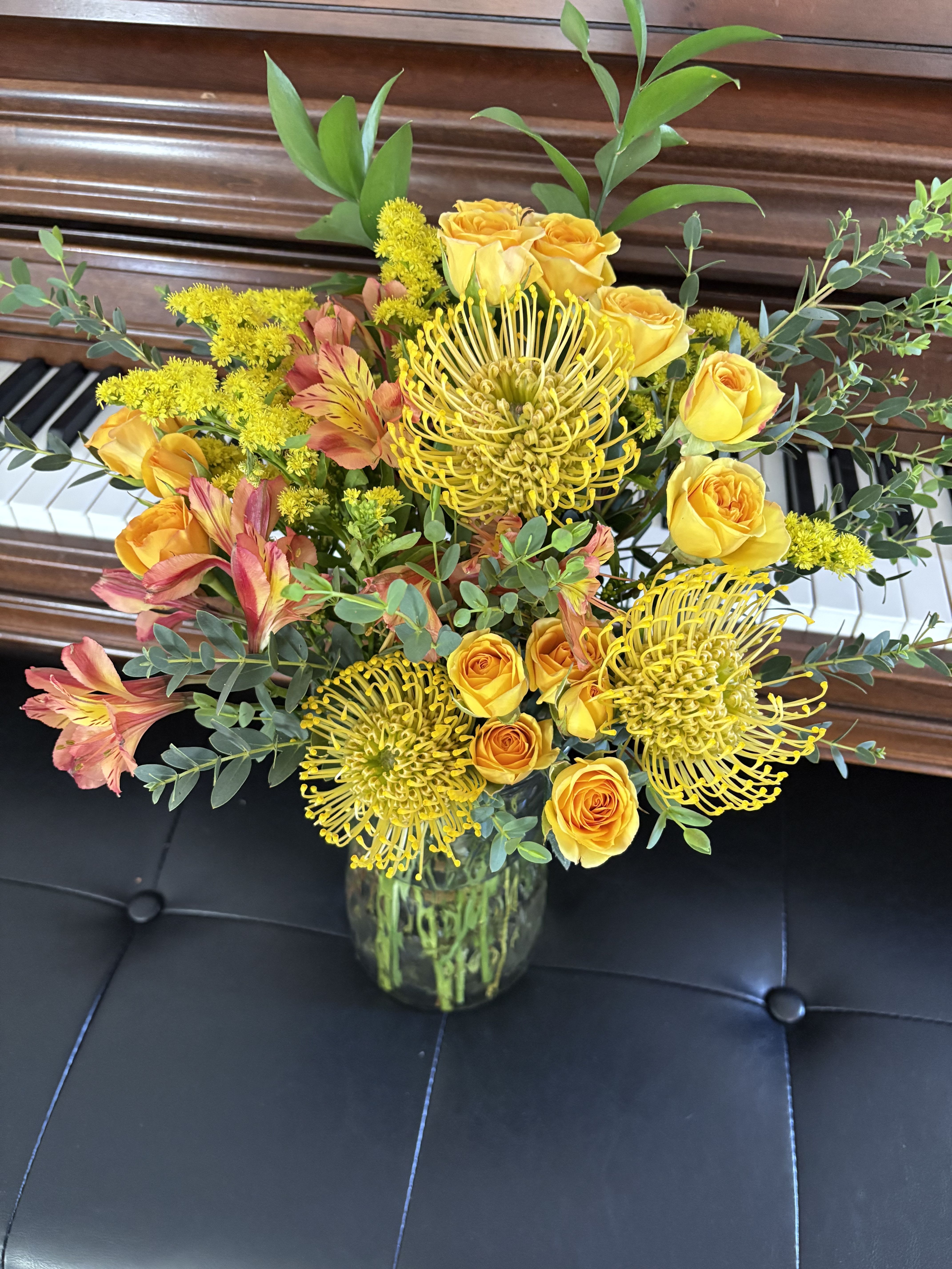 Melody in Bloom -  A vibrant floral arrangement featuring a lively bouquet of bright yellow roses, pincushion proteas, yellow solidago, and orange alstroemeria. Complemented by lush green foliage, the flowers are elegantly presented in a clear glass vase.