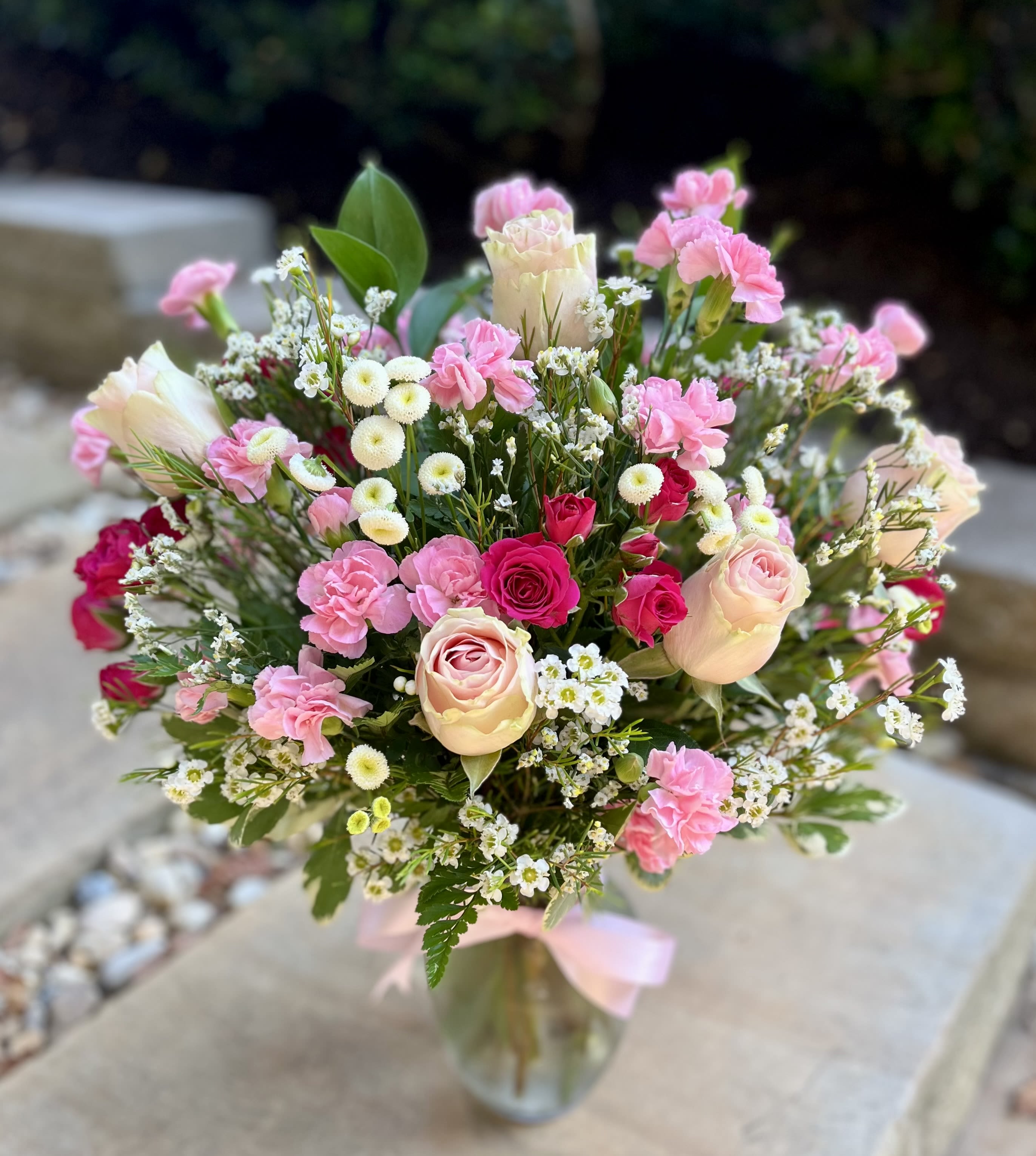 Blush Garden Delight - A charming bouquet overflowing with delicate pink roses, vibrant fuchsia spray roses, soft pink carnations, and cheerful white daisy poms. Accented with lush greenery and tied with a satin pink ribbon, this elegant arrangement evokes the serene beauty of an English garden at dawn—perfect for expressing love, gratitude, or sweet celebration.