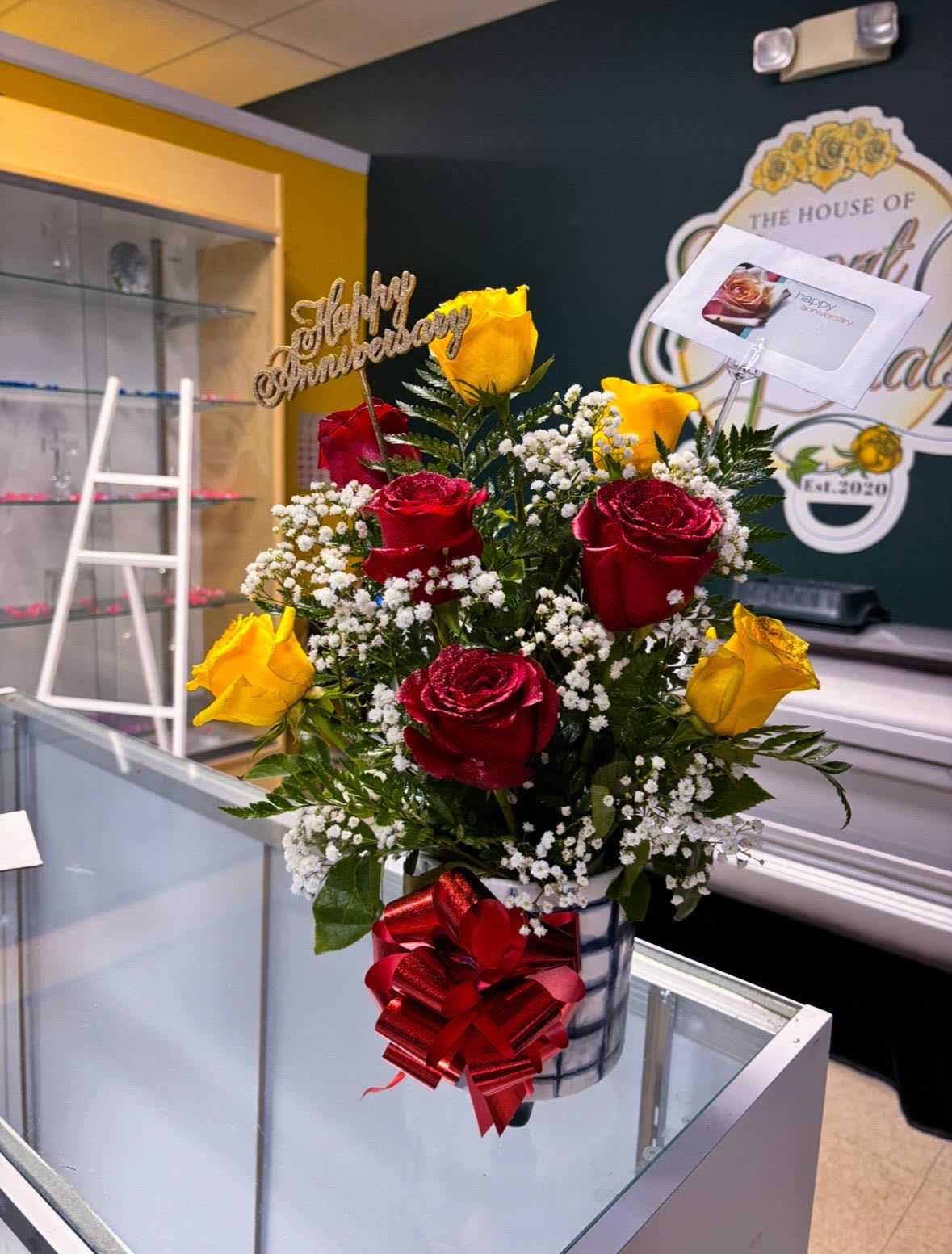  Golden Love – Anniversary Rose Vase Arrangement - Celebrate enduring love and cherished milestones with our Golden Love Anniversary Vase Arrangement. This radiant bouquet features a striking blend of classic red and sunny yellow roses—symbolizing deep passion and joyful companionship—surrounded by delicate white baby’s breath and rich greenery.  Arranged in a modern plaid ceramic vase and finished with a glittery red bow and elegant Happy Anniversary pick, this piece is perfect for romantic gestures, vow renewals, or milestone moments that deserve something truly beautiful.  Details:  Includes: Premium red roses, yellow roses, baby’s breath, assorted greenery  Vase: Stylish black-and-white plaid ceramic vase  Accents: Red glitter bow, “Happy Anniversary” pick, and custom message card  Height: Approx. 18–22”  Ideal for: Anniversaries, engagements, romantic surprises  