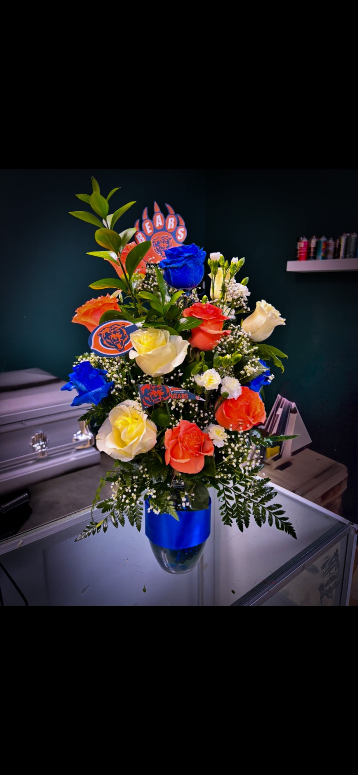 Chicago Bears Spirit Rose Arrangement - Show your team pride in full bloom! This bold Chicago Bears–inspired arrangement features a striking mix of royal blue, orange, and ivory roses, accented with fresh greenery, baby’s breath, and custom Chicago Bears keepsake picks. Designed in a sleek glass vase with a coordinating blue wrap, this arrangement is perfect for game days, birthdays, tailgates, offices, or gifts for the ultimate Bears fan.  Each piece is handcrafted with premium fresh flowers and arranged to make a statement while celebrating team spirit in an elegant way.  Flowers may vary slightly based on seasonal availability while maintaining the same look and color palette.