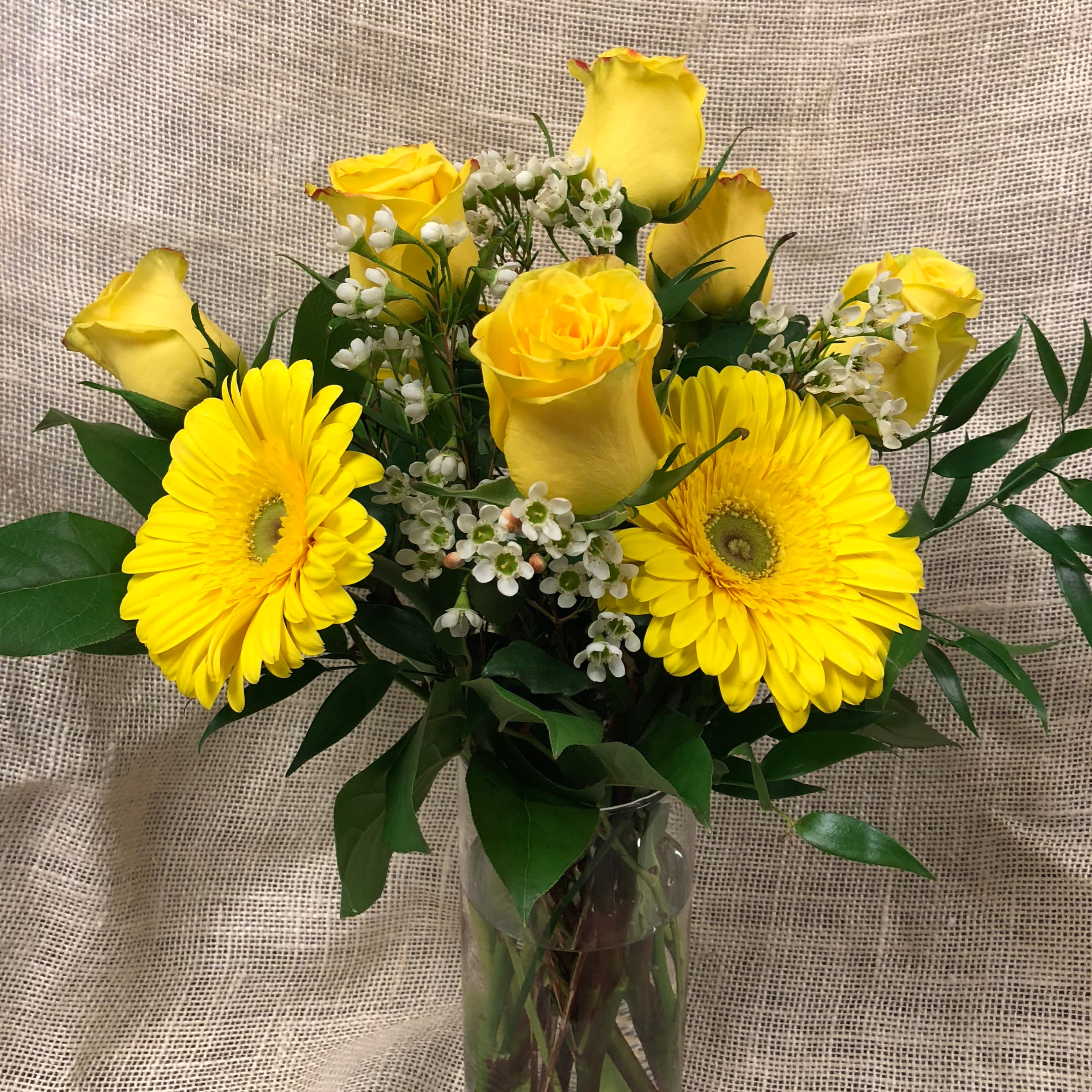 Sunshine on a Cloudy Day - Roses, Daisies - -Mother's Day Feature- This arrangement is sure to bring sunshine to someone's dark days-. Six yellow roses and three brightly colored gerbera daisies are complemented with greens and accompanying accents. Select the Deluxe or Premium upgrades for additional blossoms.  Gerbera daisies will vary, in bright complementary colors.  We custom design each arrangement for a unique, one of a kind look and feel- but be assured your design will have the same look and value as what you have ordered. Vase and color of flowers may vary.