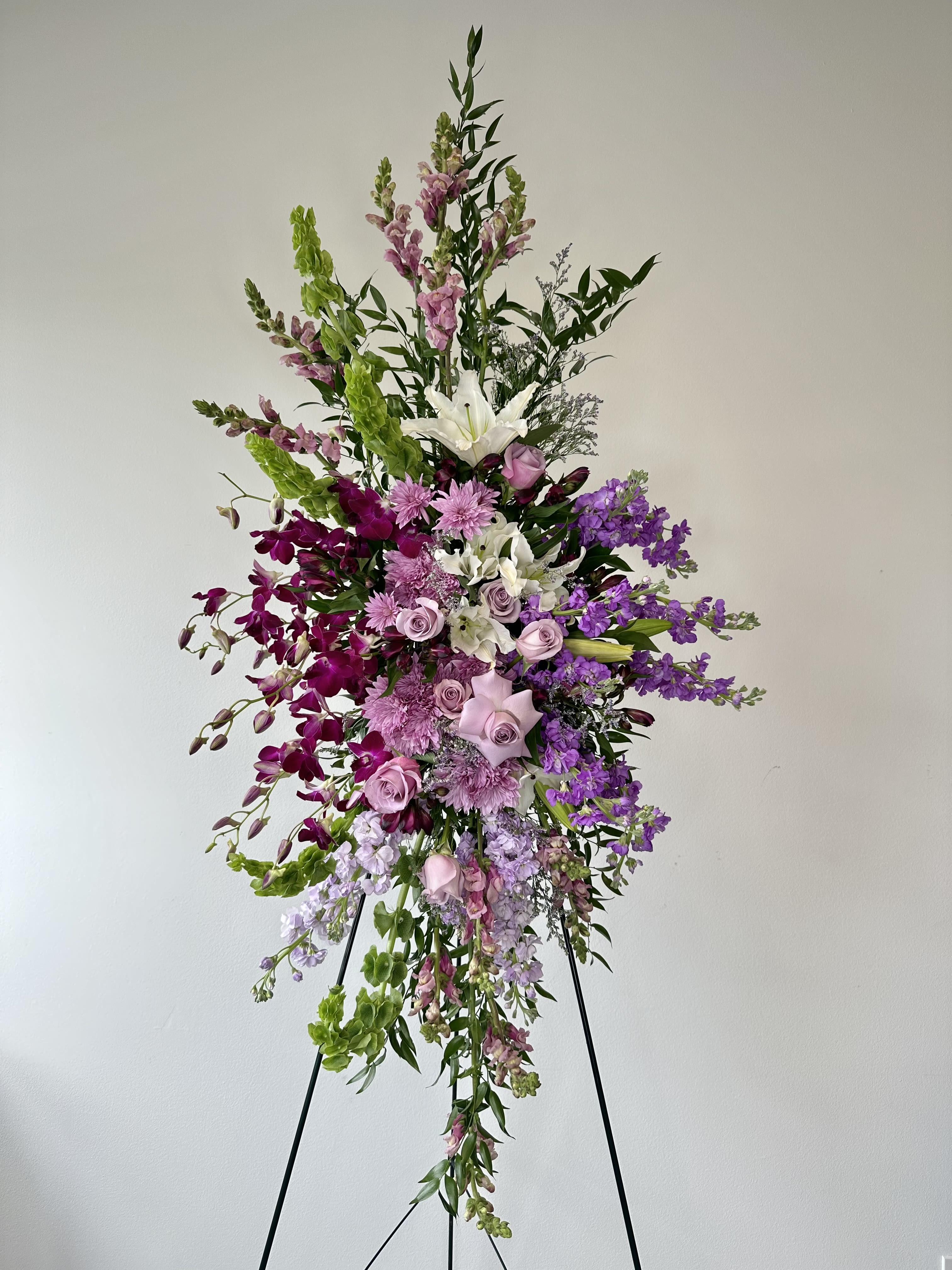 Sacred Garden Spray - A blooming embrace for the tenderhearted. The Sacred Garden Spray from Irvine Village Flowers is a floral whisper of compassion, hope, and grace, crafted to share your deepest sympathy. This lovely lavender spray weaves together a serene tapestry of purple and white blossoms—hydrangeas, orchids, roses, oriental lilies, and more—the standard standing tall at approximately 22" W x 50" H and deluxe at 30" W x 60" H. A one-sided tribute, it’s a gentle way to honor a life with beauty and peace.  Each flower offers its own quiet comfort: lavender hydrangeas spill over with heartfelt abundance and gratitude, their soft clusters a soothing balm. Pure white orchids lift spirits with their elegance, symbolizing innocence and eternal love. Roses in gentle hues breathe reverence and care, while oriental lilies unfurl with a fragrance of hope and serenity. Together, they create a sacred garden in bloom—a tribute that feels like a warm, floral hug for those grieving.  Visit Irvine Village Flowers online or in-store to send this compassionate spray, delivered fresh to wrap your loved ones in nature’s embrace. It’s a simple, beautiful way to say you’re there, sharing hope and love through every petal.