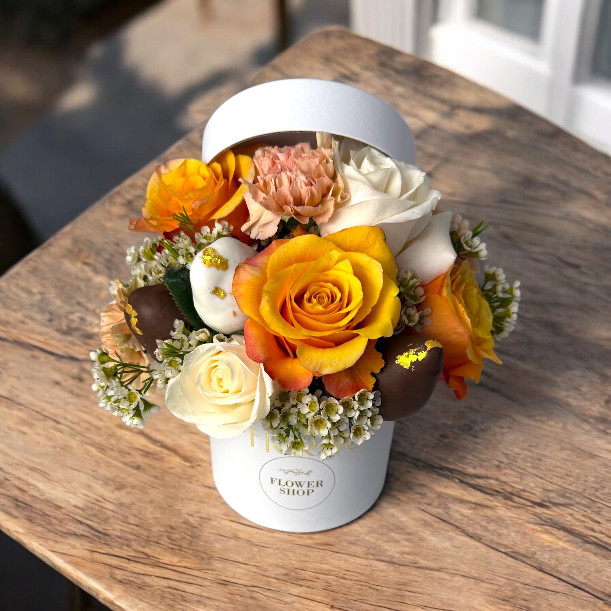 Golden Blush - Golden Blush is a floral gem that radiates warmth and elegance. Soft golden roses, coral carnations, and delicate white wax flowers are paired with individually coated white and dark chocolate-covered strawberries, adorned with edible gold leaf. All beautifully presented in a small round white box — a refined gift full of sweetness and charm.