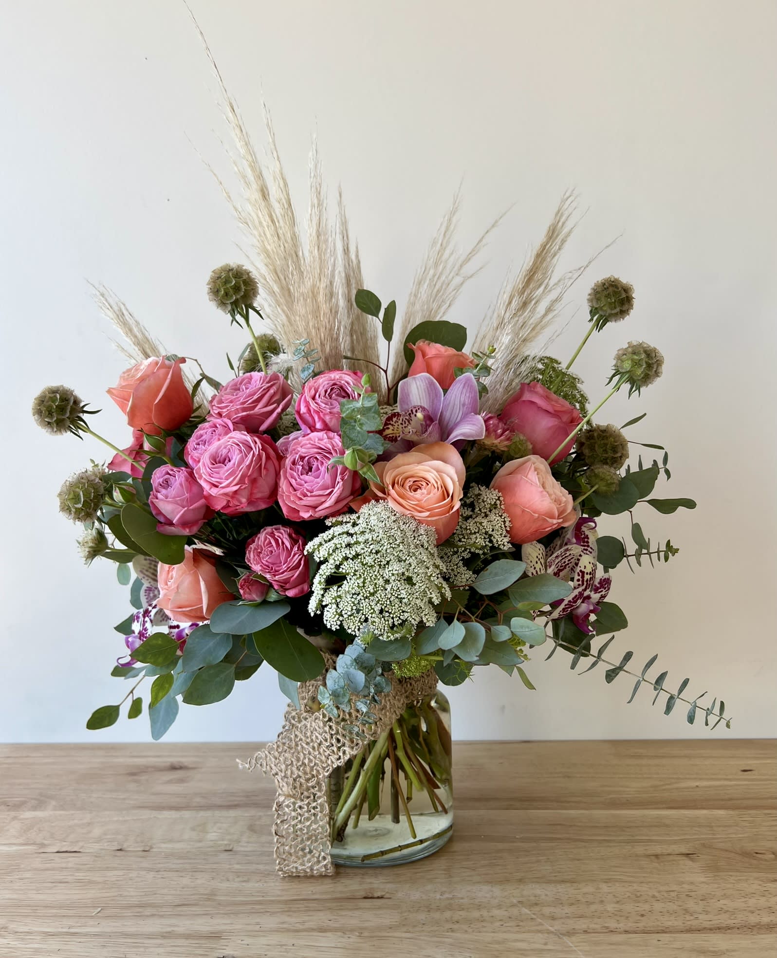 Beach Vibe - Bring the serene elegance of the coast indoors with this unique beach-inspired floral creation.   Artfully arranged in a clear glass cylinder wrapped with a natural tan burlap ribbon, this design beautifully balances rustic texture with refined blooms.  Displaying lush garden roses, exotic cymbidium &amp; phalaenopsis orchids, accented with airy green mist and feathery pampas grass. While scabiosa pods and lush eucalyptus add organic depth &amp; movement.  Perfect for coastal weddings, summer celebrations, or anyone dreaming of salty air and endless horizons, this arrangement captures the effortless beauty of the beach with a sophisticated twist.