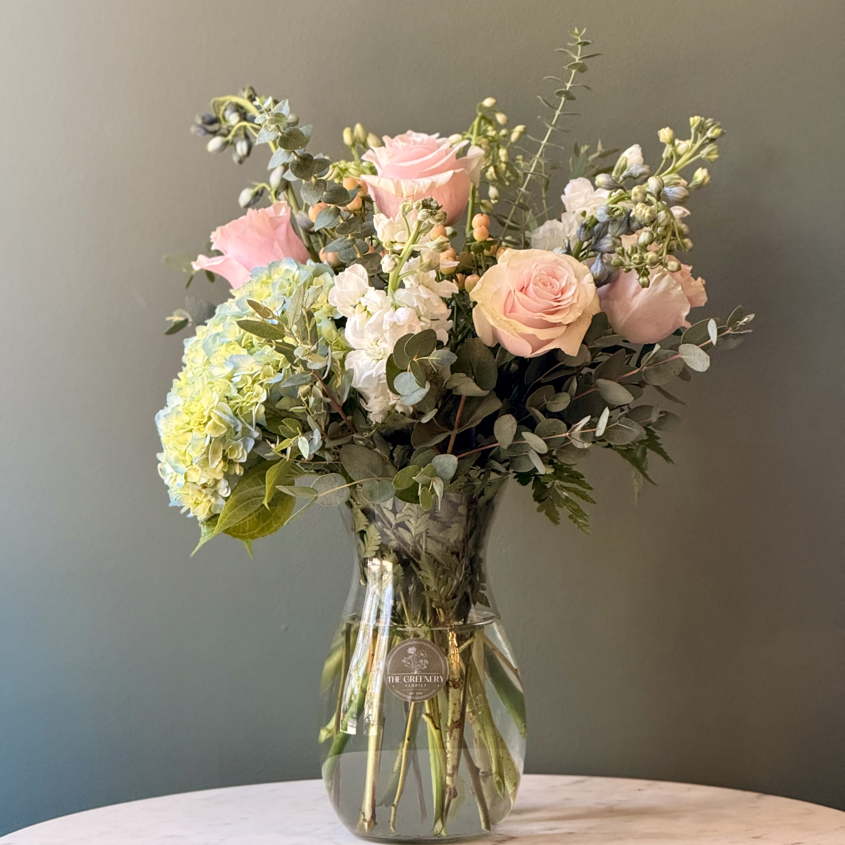 Lowcountry Dreamin' - A soft, elegant blend of pastel blooms. Featuring: - Ecuadorian Roses - Blue Hydrangeas - White Delphinium - Light Blue Delphinium - Hypericum - Stock - Styled with premium seasonal foliage  Presentation Options: Tall clear vase measuring approximately 8"/9" H. (vase included in price) Every order includes: A custom message card - simply enter your message at checkout and we'll take care of the rest. Please note: Flowers are subject to seasonal availability. If a listed flower is unavailable, our designers will substitute it with flowers of a similar shape, color, and style to maintain the overall aesthetic.