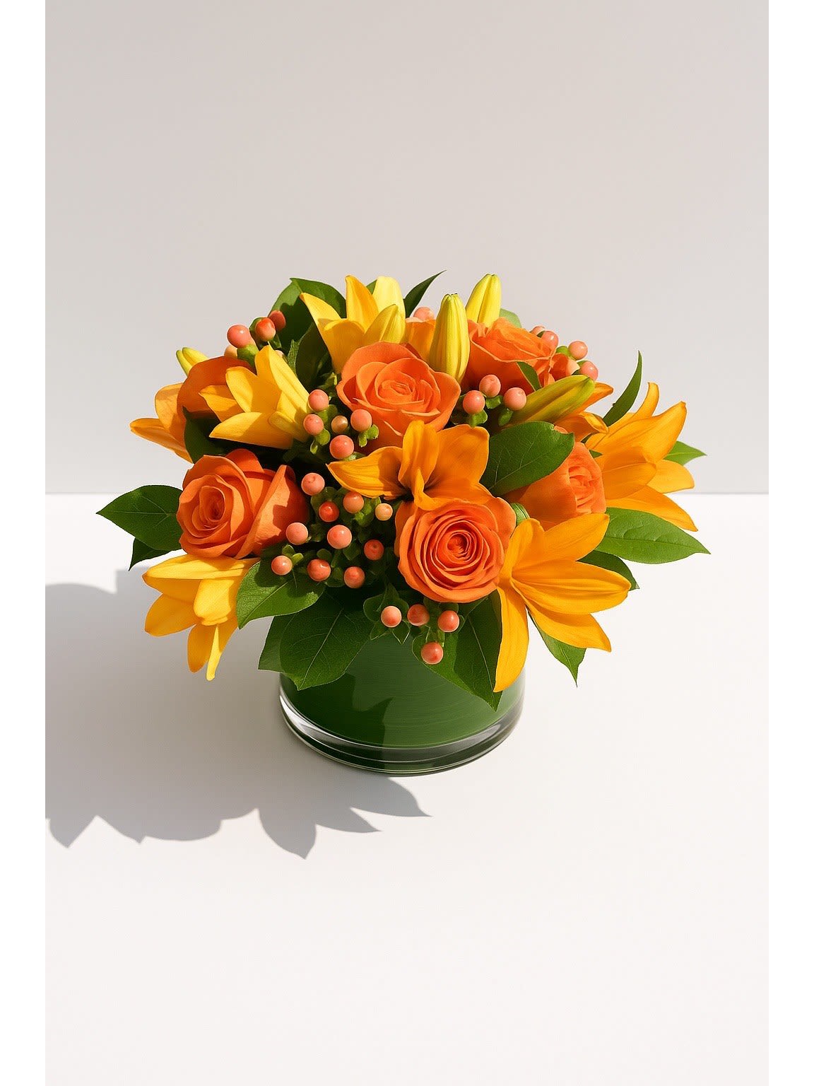 Tangerine Twist  - A burst of vibrant warmth in every petal. Tangerine Twist features bold orange roses and lilies paired with soft greenery and berry accents, creating a lively arrangement that radiates joy. Designed in a sleek glass cylinder wrapped with lush foliage, this piece brings an instant pop of sunshine to any space — perfect for birthdays, celebrations, or brightening someone’s day.  Style: Modern &amp; Lush Color Palette: Tangerine, Coral, and Warm Green Approximate Size: 10–12” wide