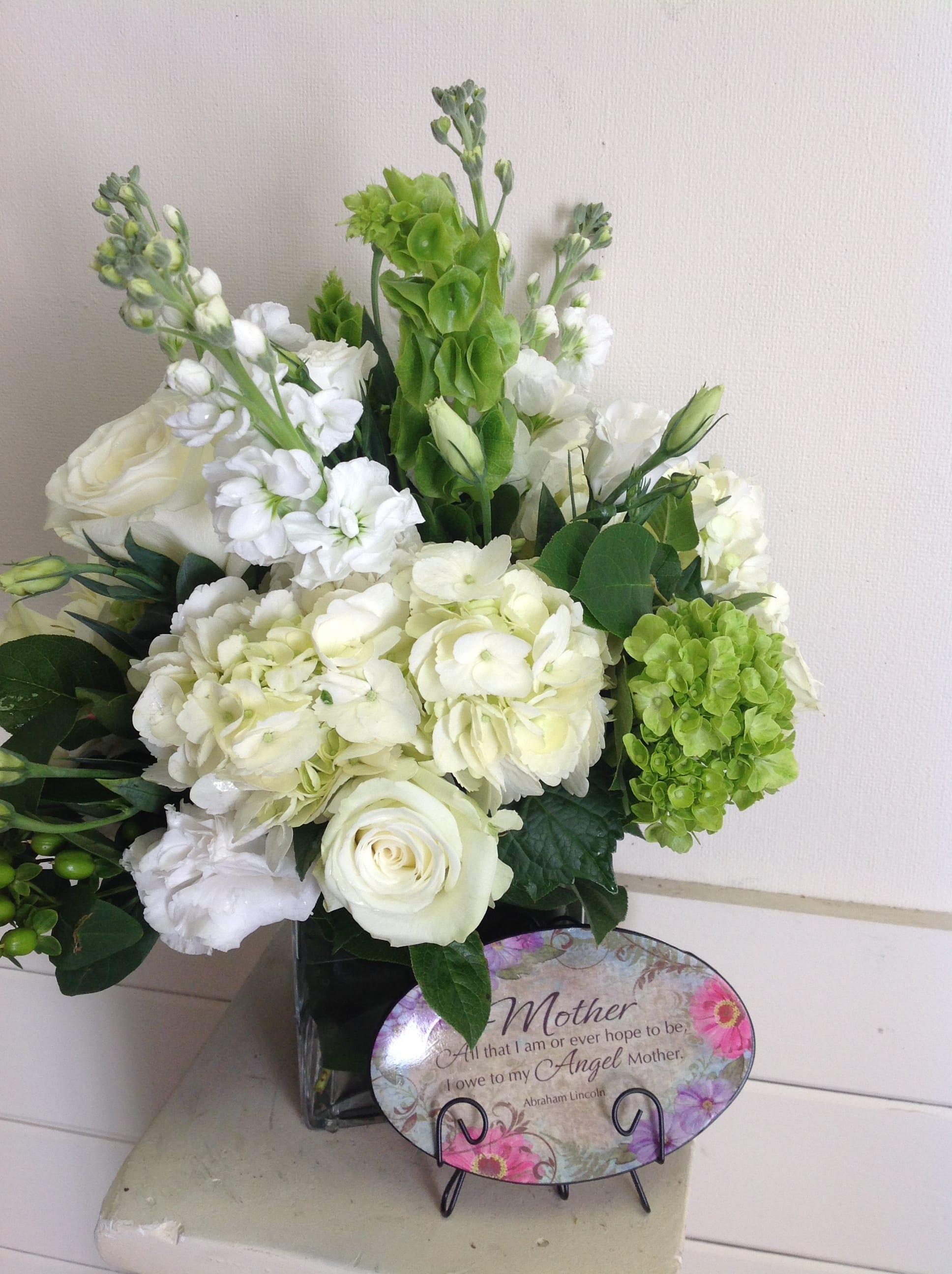 Motherly Love - 6 inch clear cube filled with white roses, white stock. white hydrangea, white lisianthus, green bells of ireland and green hydrangea wrapped in an aspidistra leaf.