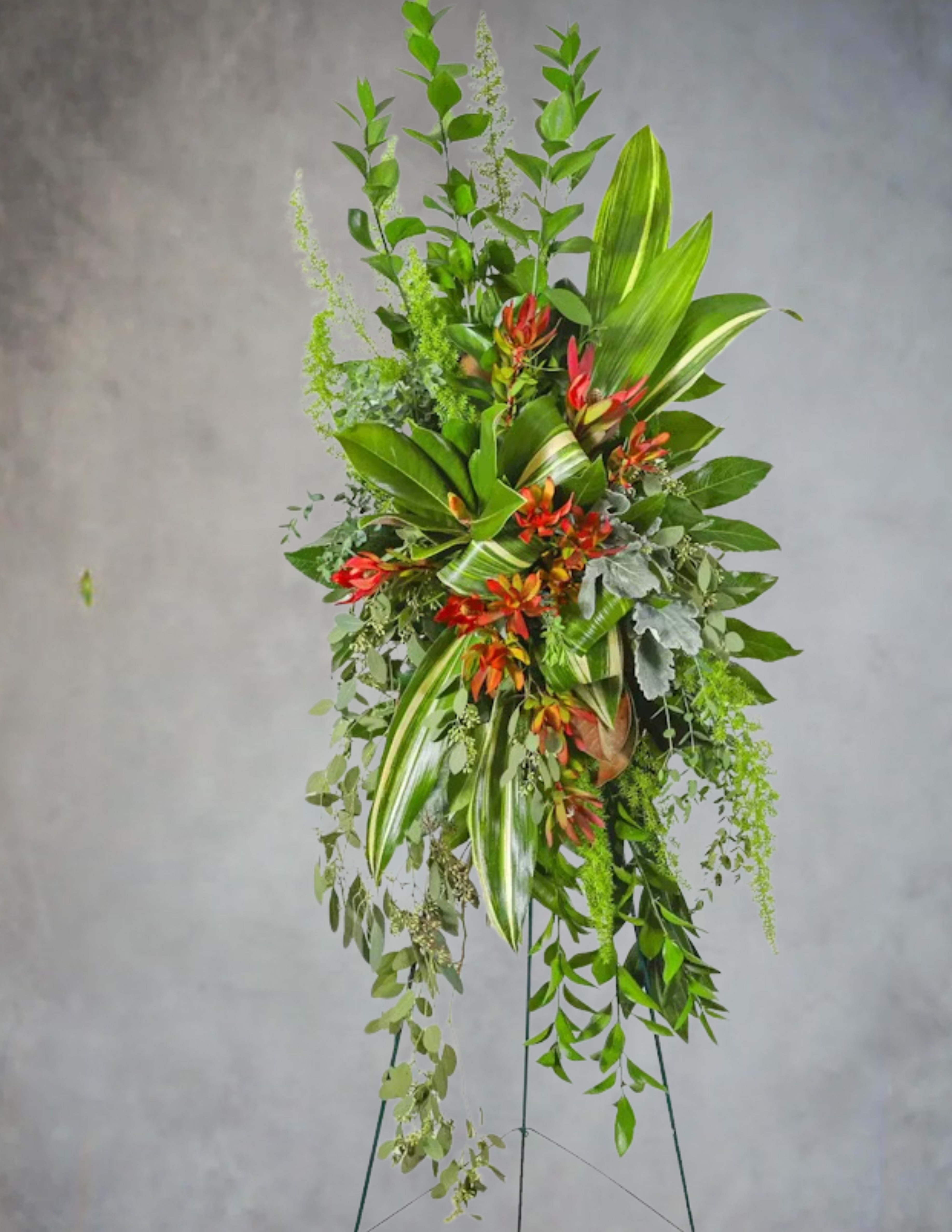 A Life Well Rooted - A Life Well Rooted  A Life Well Rooted is a graceful standing tribute that honors a legacy built on strength, depth, and quiet beauty. Lush layers of vibrant greenery create a foundation that feels grounded and enduring, while rich red‑orange blooms rise through the foliage like moments of passion, joy, and color that defined a well‑lived life. The asymmetrical, garden‑inspired design evokes the feeling of a flourishing landscape—one shaped by resilience, generosity, and growth.  This arrangement offers a heartfelt expression of remembrance, celebrating a life that took root, reached outward, and left the world more beautiful.