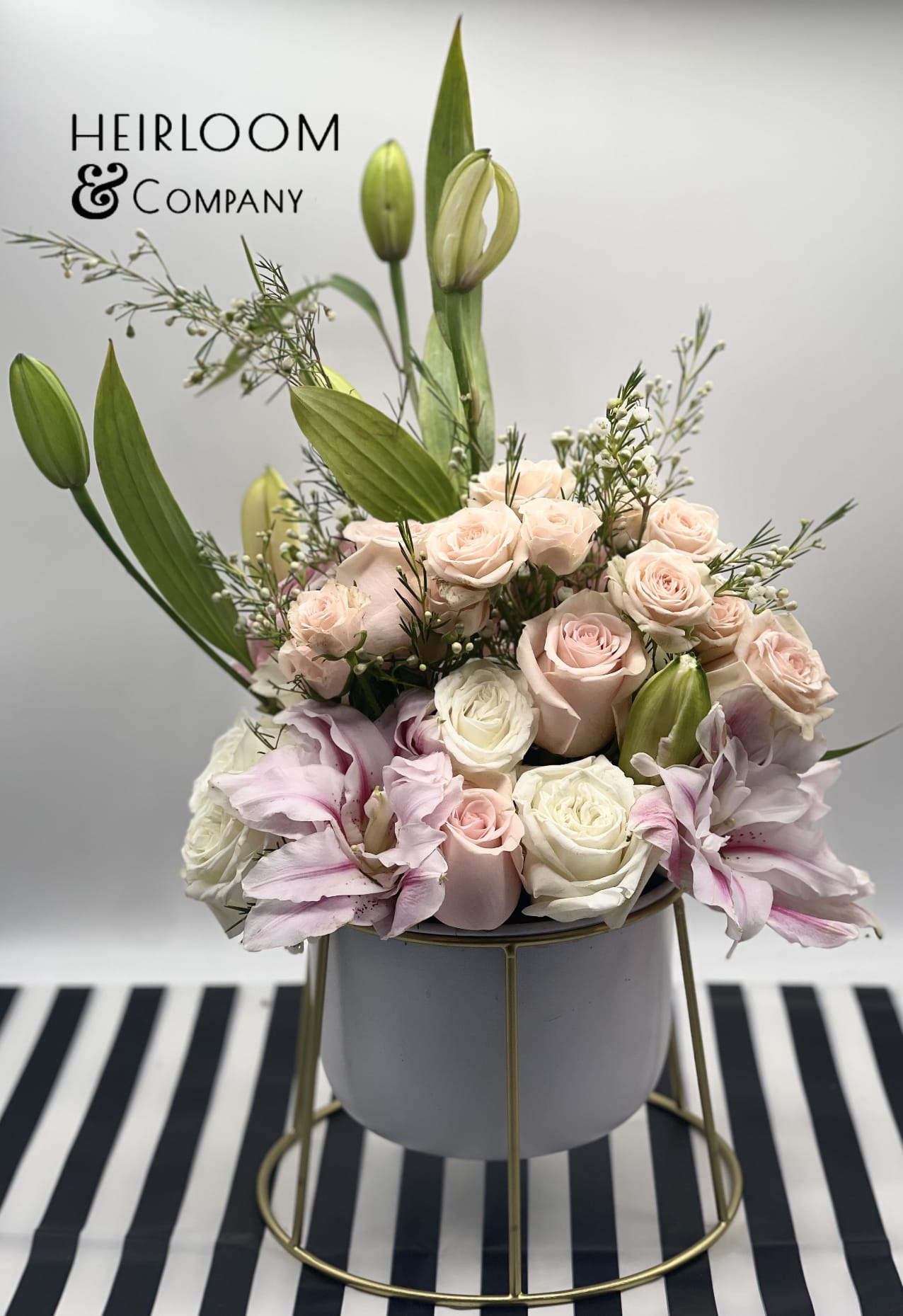Lilly wildflower - A tall wildflower arrangement in a white container floating on a golden rack, filled with lilies, garden roses, mini spray roses, in shades of pink and white.