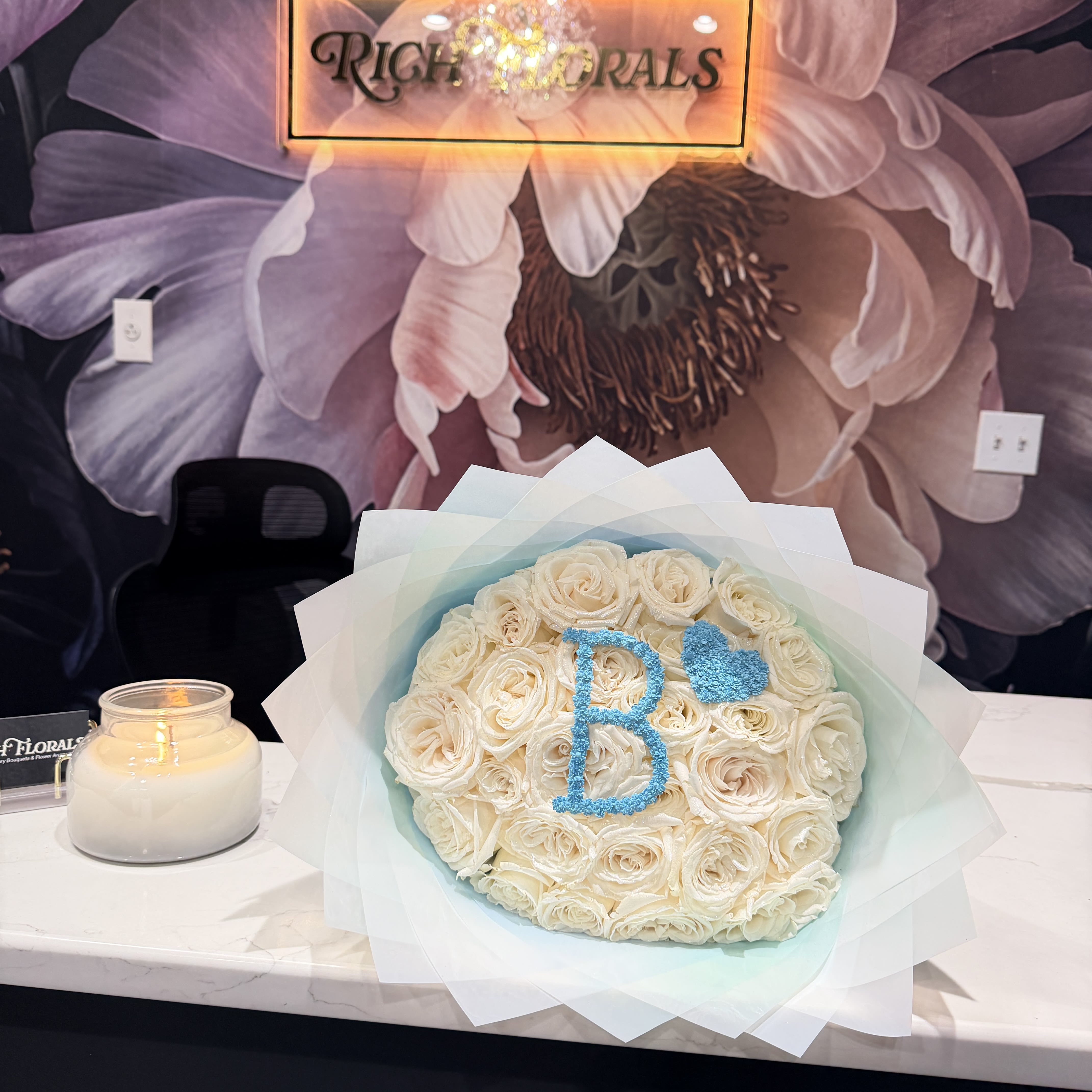 Baby Boy Bouquet - White Roses, Blue Baby’s Breath "B" &amp; Heart  Say it in roses… but make it baby blue. Our Baby Boy Bouquet is what happens when soft meets statement. A full, lush dome of premium white roses wrapped in light blue paper and finished with a subtle silver sparkle dust that catches the light just right. Clean. Polished. Unforgettable. Front and center? A custom blue baby’s breath “B” and heart, giving it that personalized touch that feels thoughtful without trying too hard. It’s a “welcome to the world, little king” moment.  Whether you’re celebrating a new baby boy, styling a baby shower, surprising a new mom at the hospital, or sending love from afar, this bouquet speaks before you do. Because let’s be honest… grocery store flowers could never.  Perfect For: - New baby boy gifts - Baby shower centerpieces - Hospital flower delivery for new moms - Welcome home baby celebrations  Each arrangement is handcrafted using premium roses sourced specifically for your order. No pre-made inventory. No shortcuts. Just fresh blooms designed for your moment.