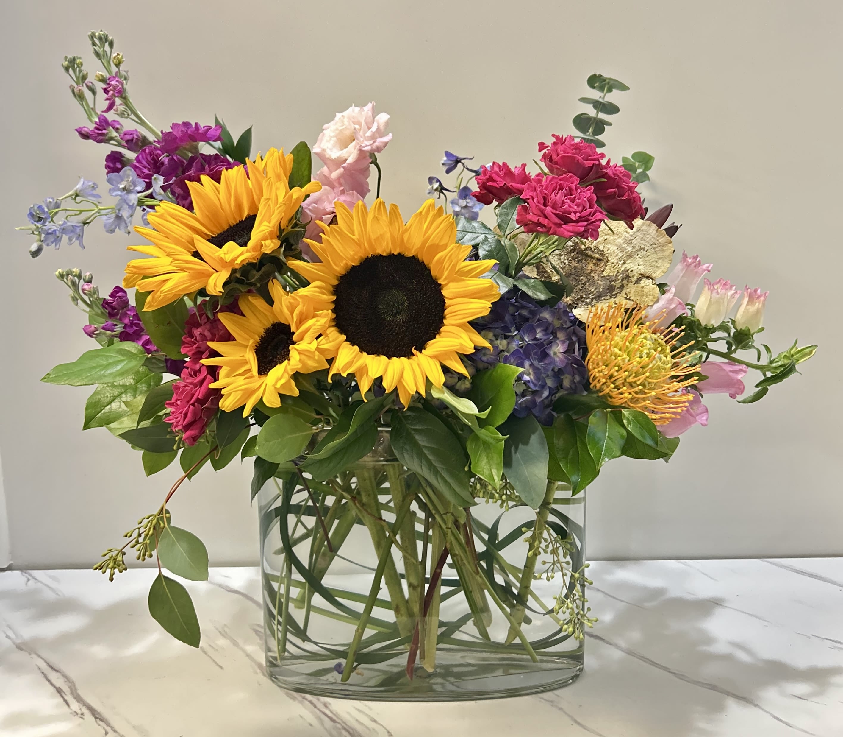 Sunny-Licious  - Sunny-Licious is a vibrant and upscale floral arrangement that brings sunshine to any occasion. Featuring bold sunflowers alongside a mix of other lively, colorful blooms, this design radiates warmth and cheer. Its modern, sophisticated look makes it the perfect choice for those seeking to brighten a special moment with a burst of color and elegance.