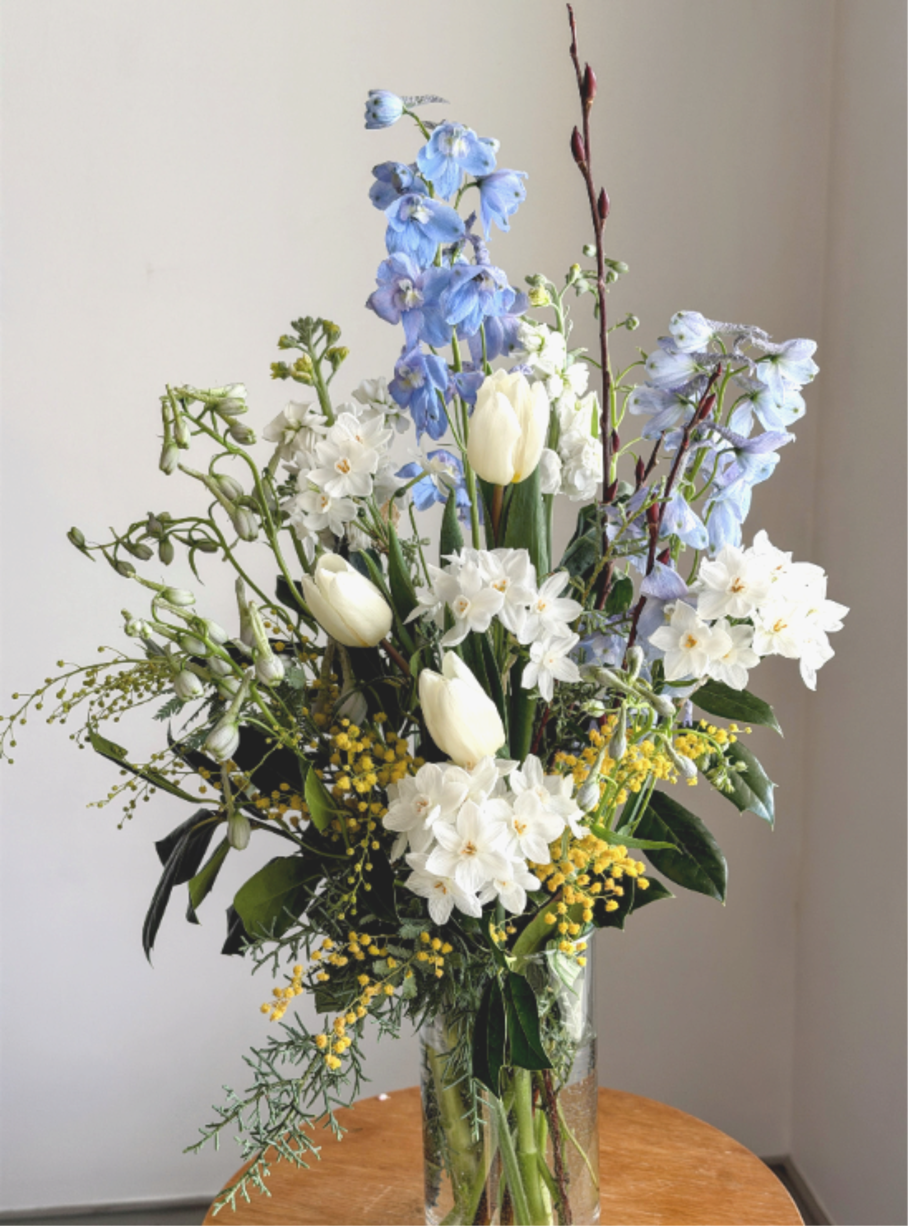 Winter Blues - Send a little pick me up with this classic blue &amp; white arrangement. Accented with a bit of yellow mimosa this cylinder vase comes with blooms such as delphinium, tulips &amp; narcissus.