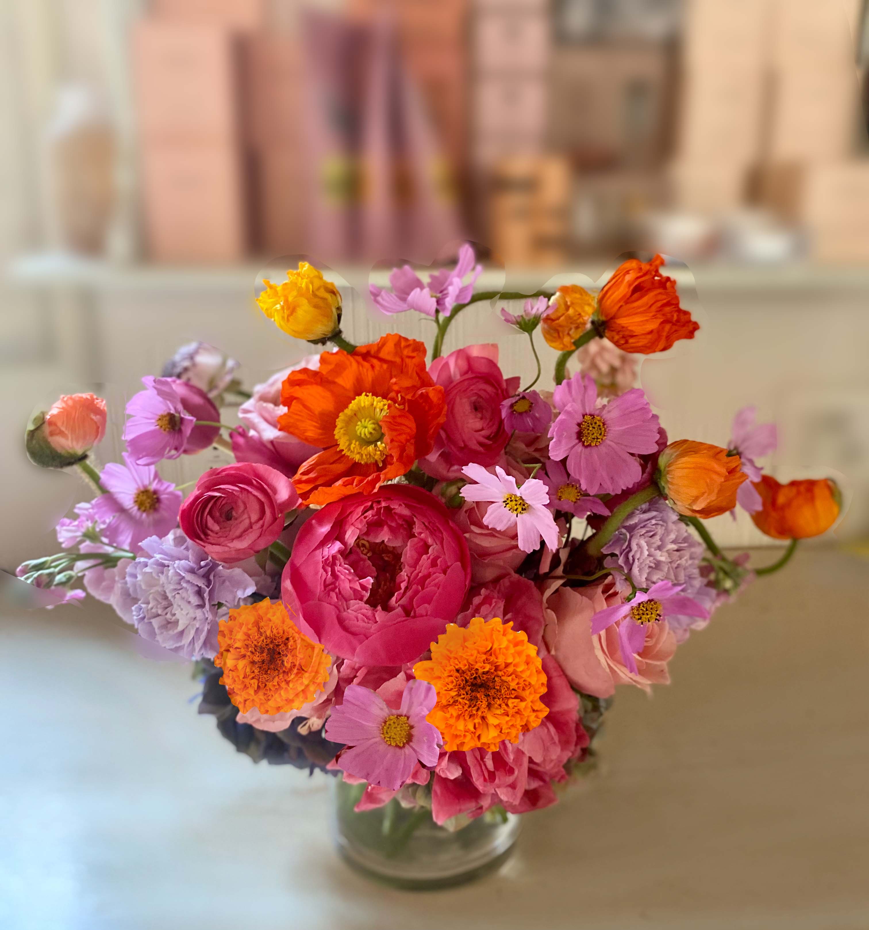 Holly Arrangement - Lively and bright Holiday arrangement of gorgeous warm and vivid colors of orange, pink, yellow and purple, of seasonal blooms like ranunculus, roses, poppies, peonies, cosmos and varying stems of each season, placed in a clear 5" by 5" vase. Choose Deluxe for more stems and a larger vase, and Premium for a large variant of the All Saints Arrangement in a larger vase with even more stems.