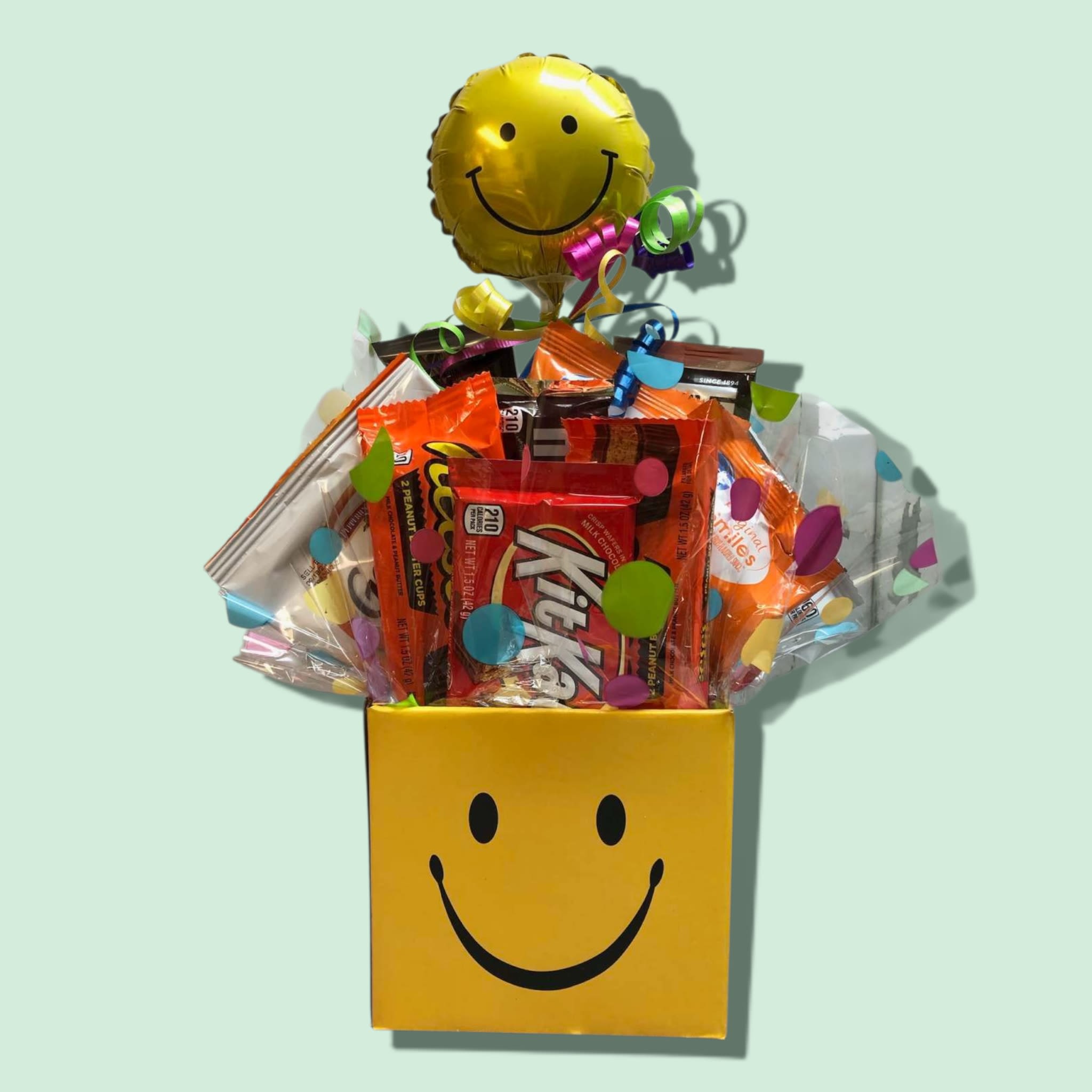 Snacky Smiles Box - A delightful gift for anyone, our Snacky Smiles Box will bring joy to all! A mixture of popular candy bars, crackers or cookies, and gummies is festively displayed in our smile box and topped with a 4" mylar stick balloon. Approximately: 6 bars, 2 crackers or cookies, and 2 gummies