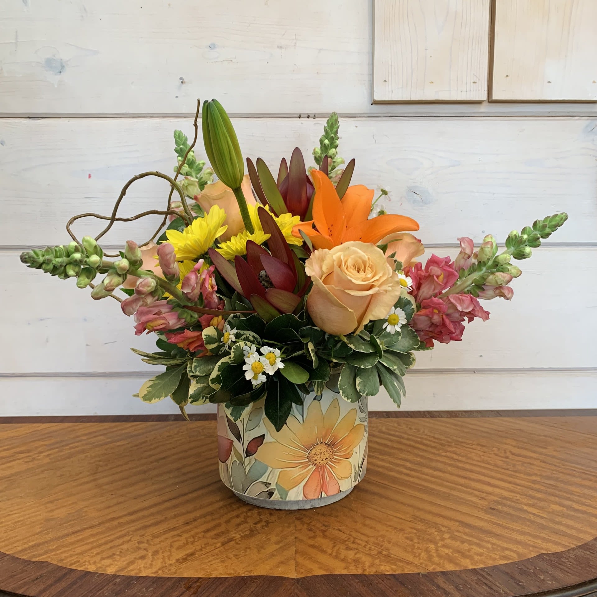 Summer's Kiss - Apricot roses and orange lilies, with orange snaps, yellow daisies, and safari sunset in a watercolor wildflower printed pot with curly willow and seasonal filler flower. Approximately 11" x 11" for the standard.