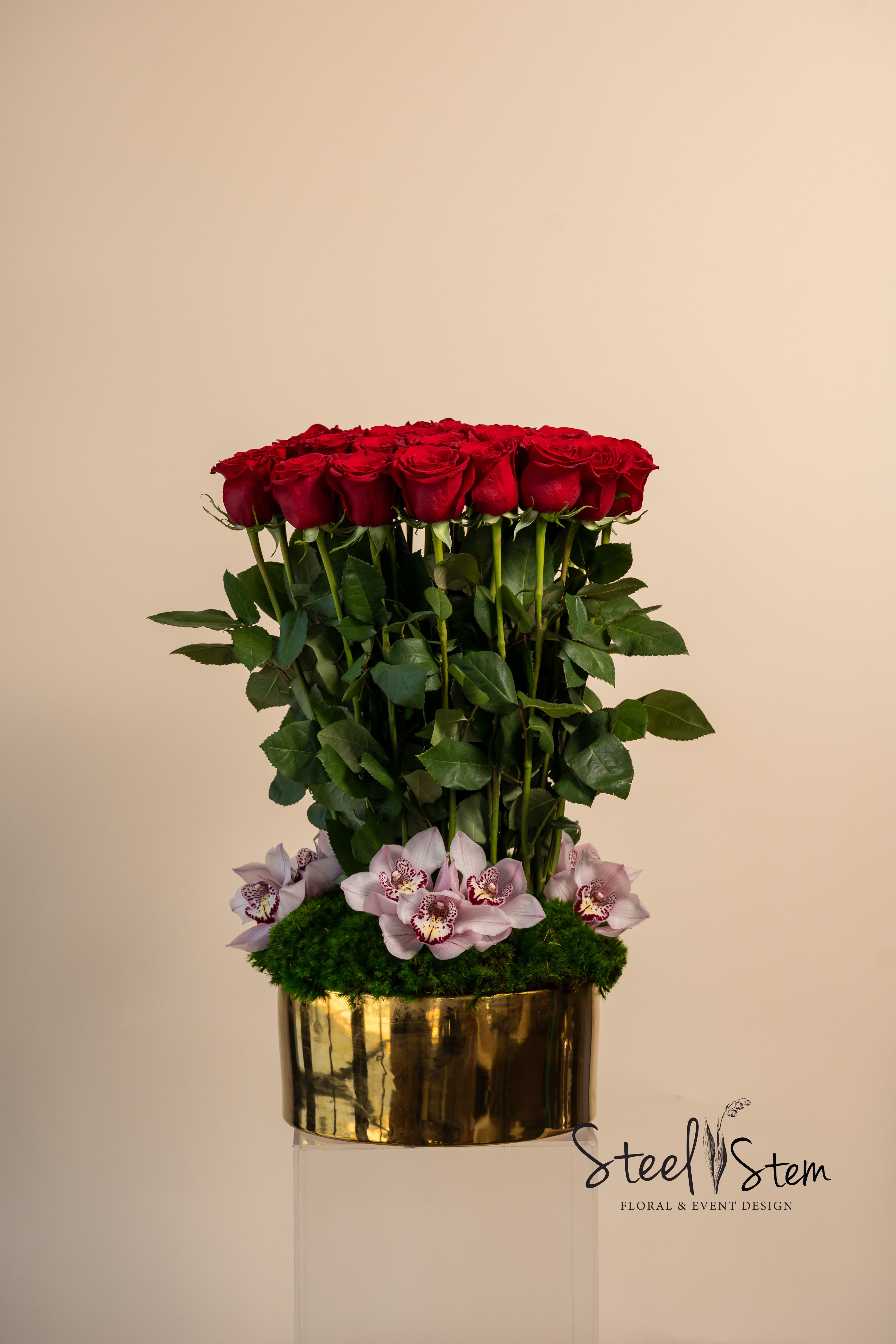 Scarlett Symphony - Scarlet Symphony  Size: Approx. 6” x 9”  A masterpiece of modern romance, 12”x20”Scarlet Symphony is a striking floral composition that blends structure, elegance, and passion. Featuring a sculptural topiary of red roses rise above a bed of green moss and  delicately accented of cymbidium orchids, in a sleek gold container    