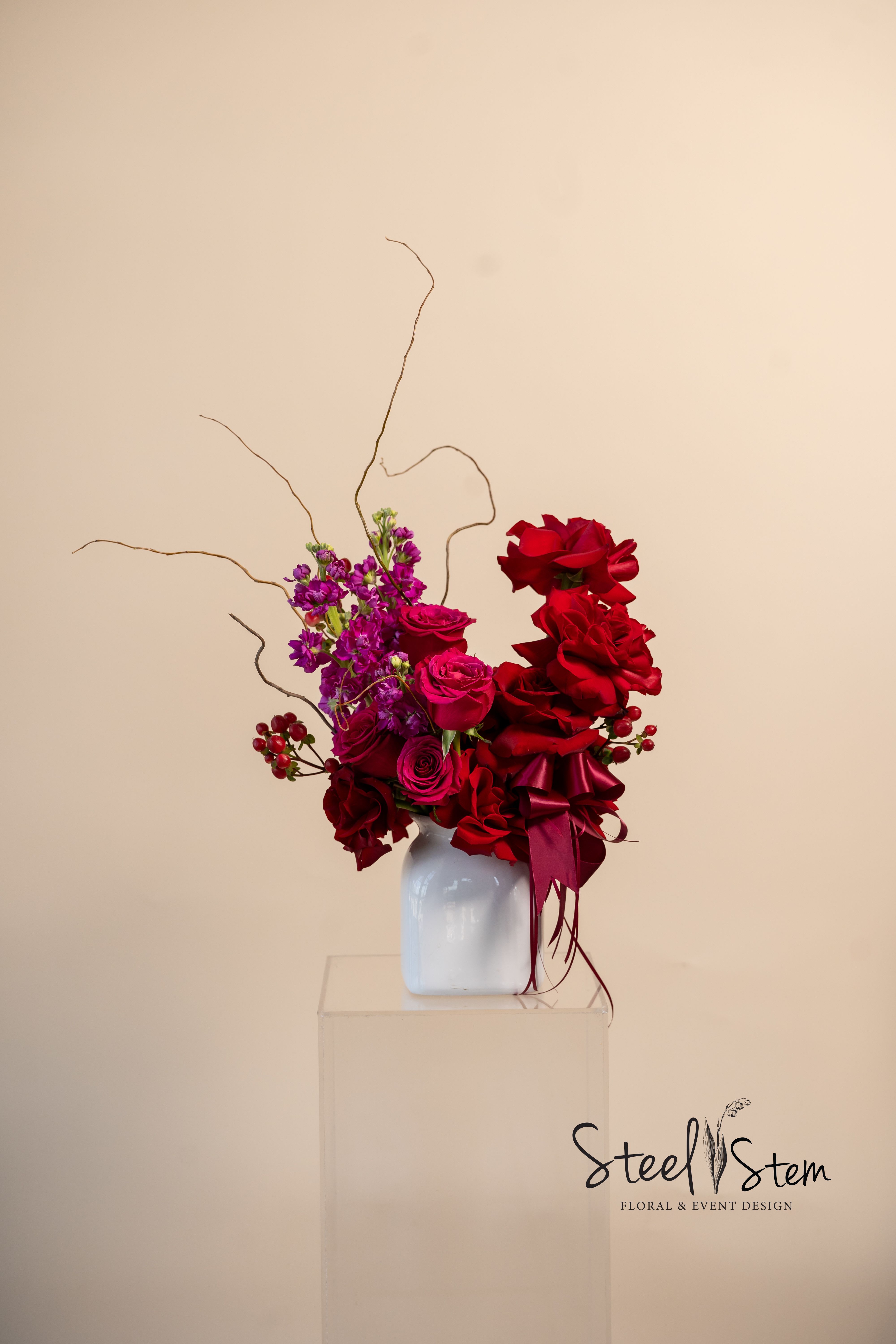 Passion Gradient  - Passion Gradient is a mesmerizing  6” x 9” floral arrangement designed to captivate, this ombré-inspired creation blends red roses, hot pink roses with fuchsia-toned stock, creating a stunning visual symphony of passion and desire. Accentuated with delicate flowing satin ribbon, and organic branches, in a classic white vase this design balances elegance with bold movement. 